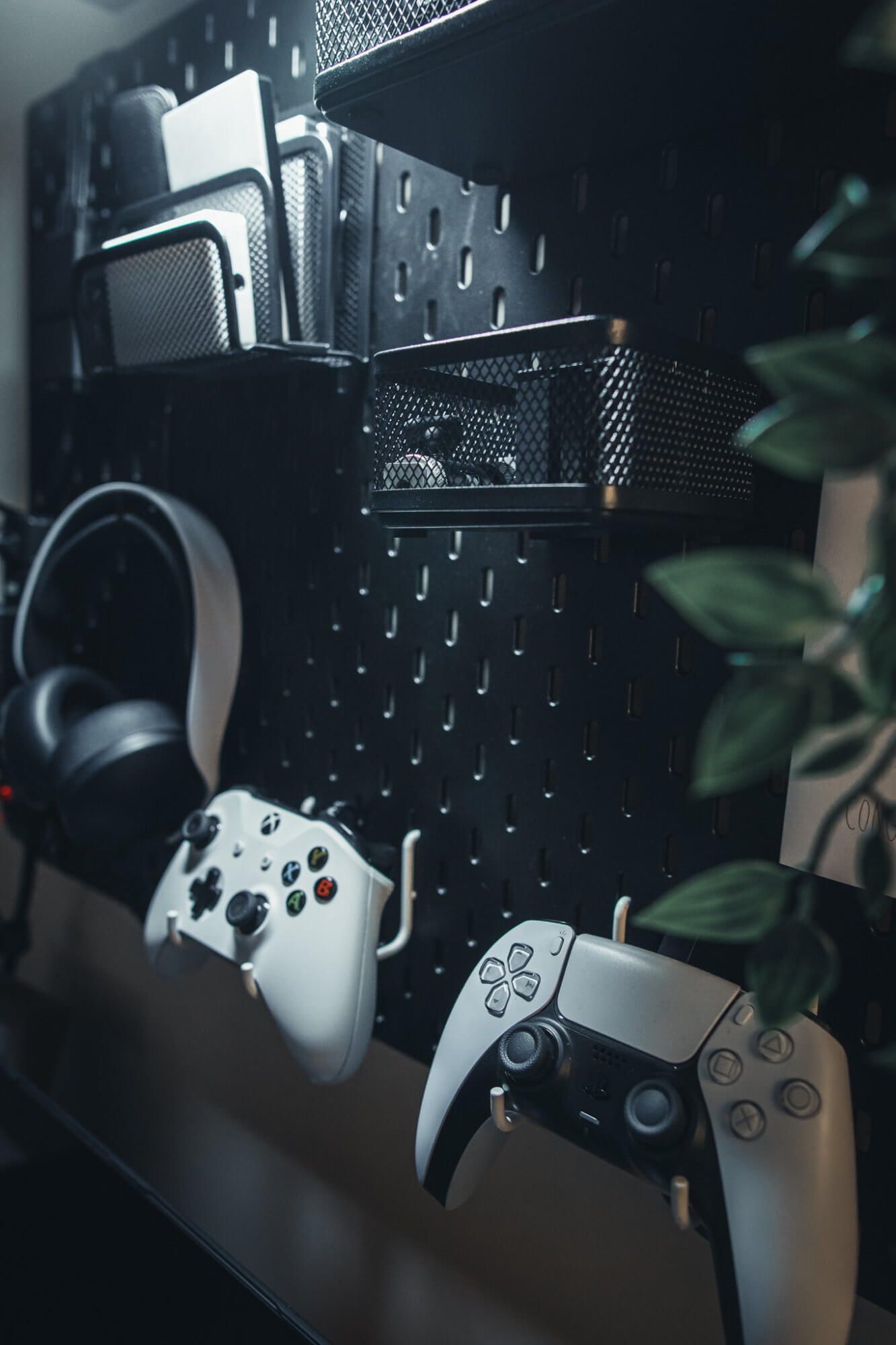 Two PlayStation controllers, a headset and other home office accessories on a black IKEA pegboard 