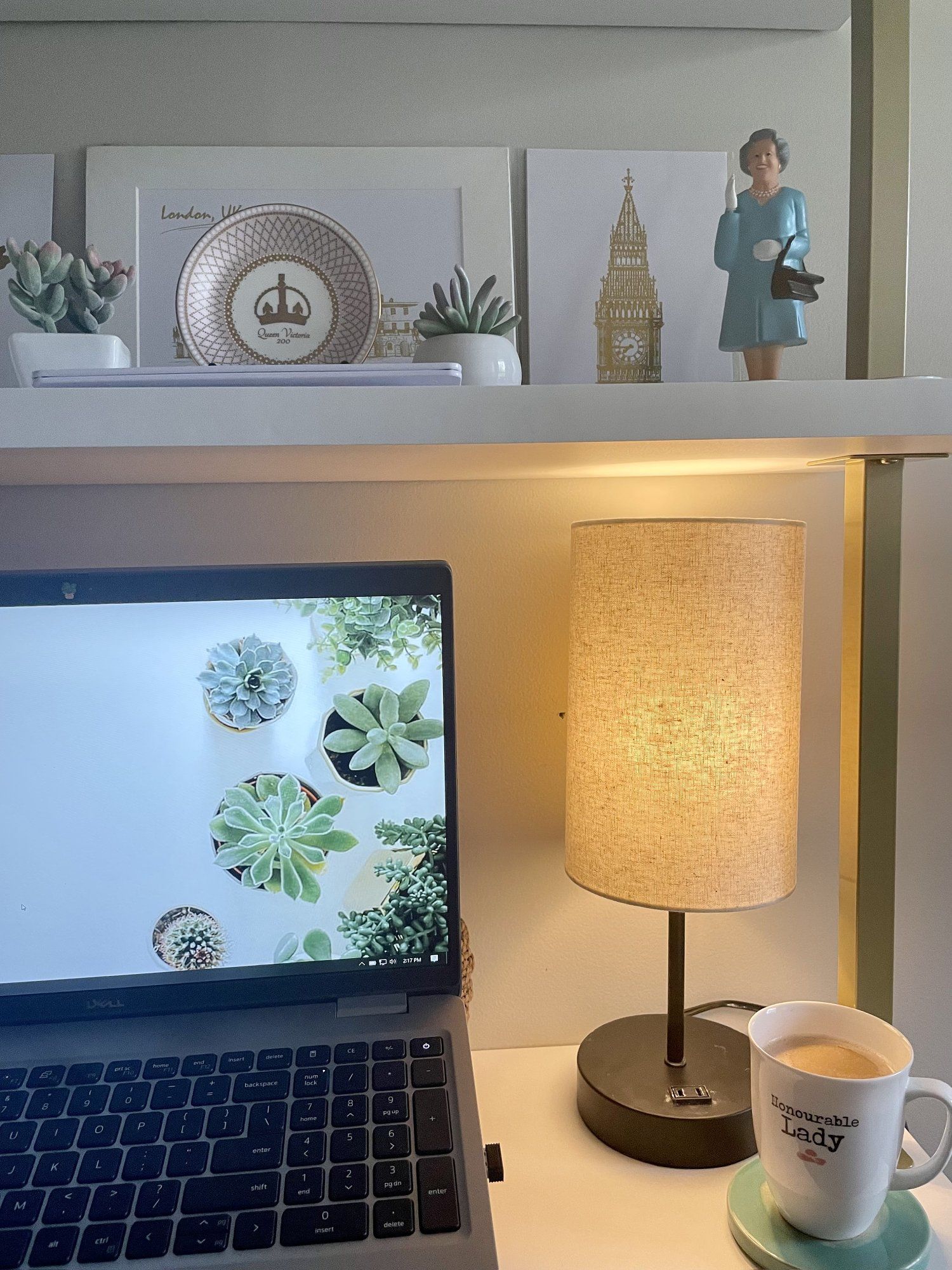 A desk setup featuring the Dell laptop, lamp, Honourable Lady mug, and some British souvenirs on the shelves