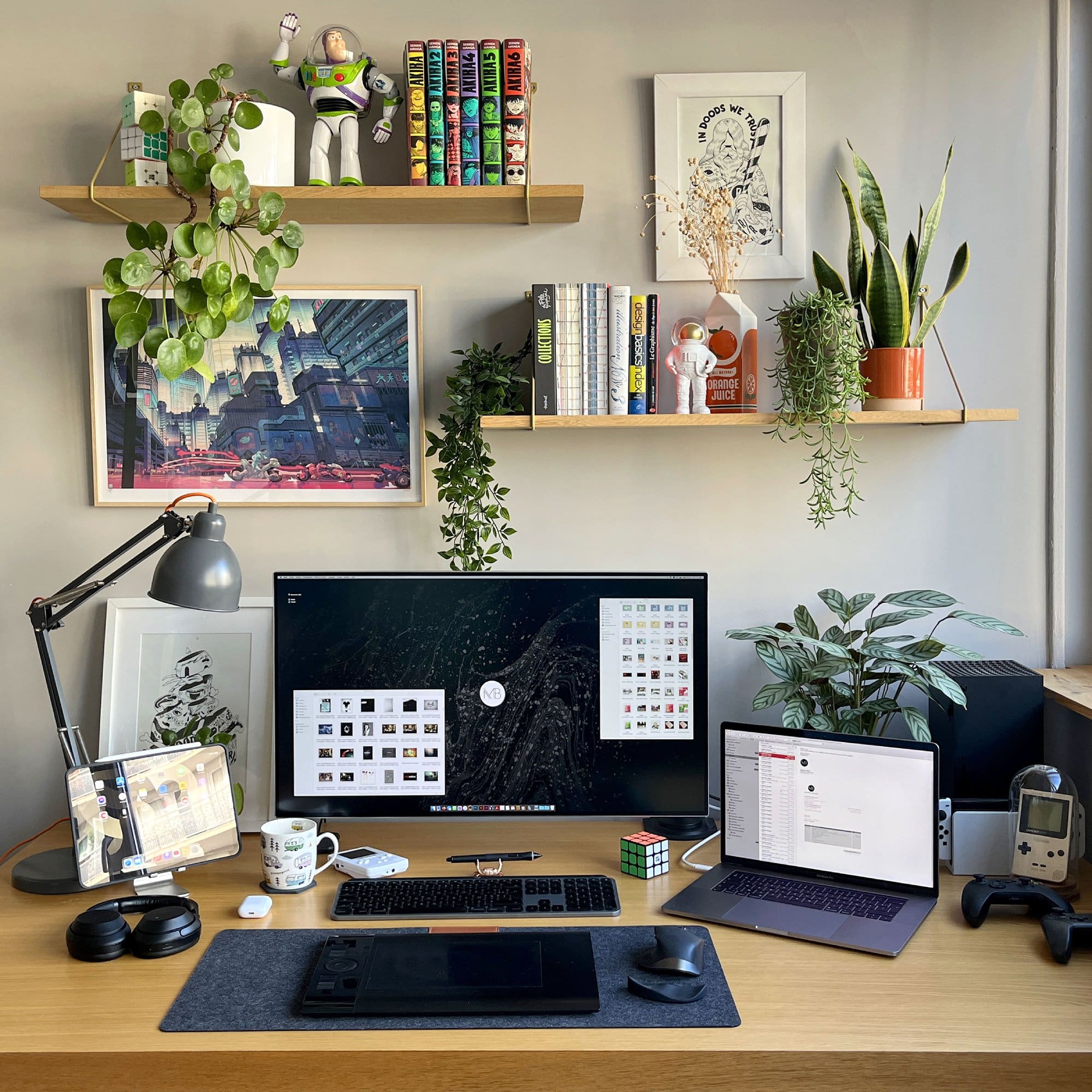 A well-organised graphic designer’s home desk setup with shelves for some plants, comics, books, and figurines on the wall