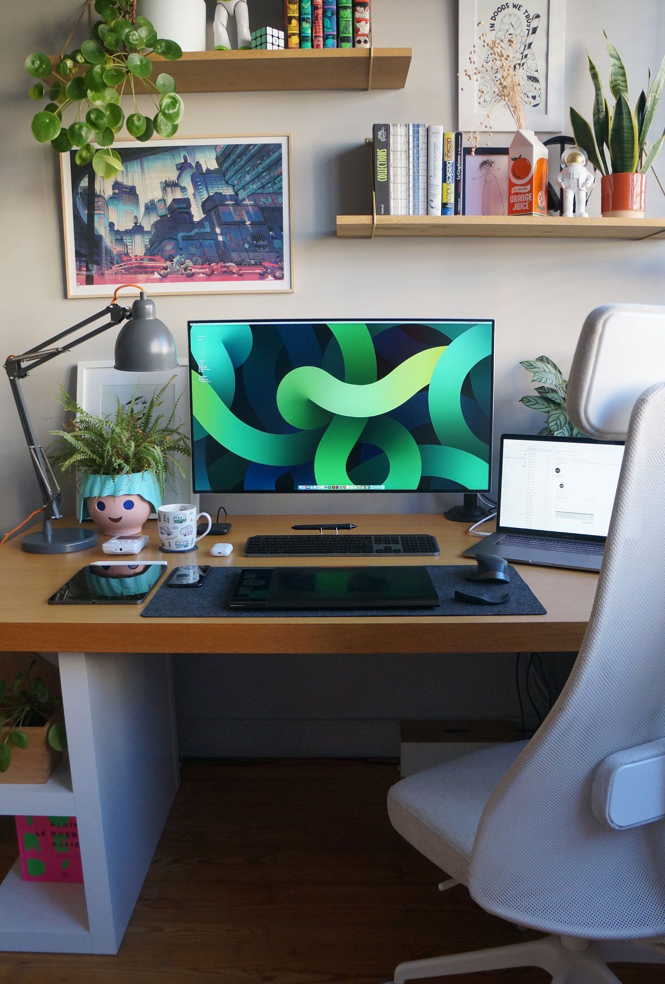 A neat and bright graphic design home office setup, featuring an LG monitor, a MacBook Pro, an IKEA JÄRVFJÄLLET office chair, bookshelves, plants, and a limited edition illustration Akira by Mathieu Bablet on the wall