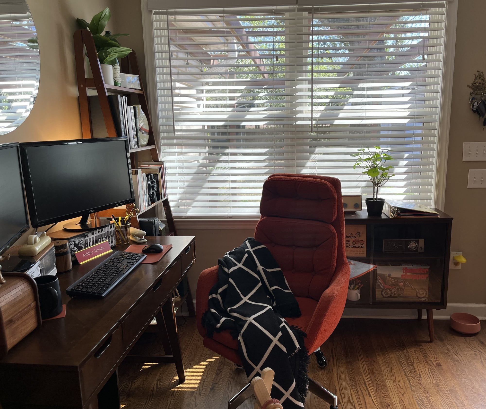 A close-up view of a mom’s home workspace showcasing a wooden desk with a multi-screen setup, a cosy vintage chair, and a large window