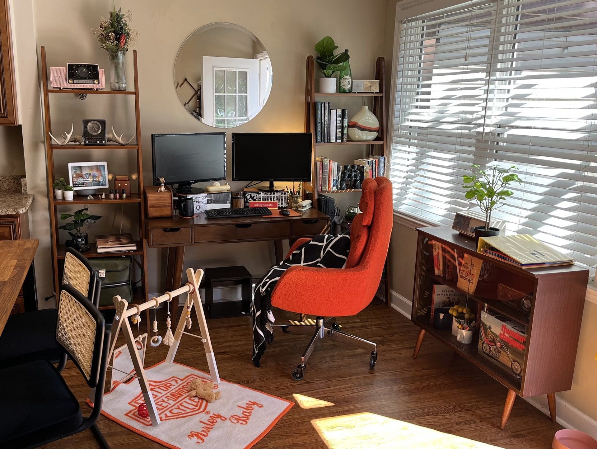 The bright and inviting working mom’s home office featuring the wooden desk with a dual-monitor setup and a cosy orange chair. The room has a large window that allows for plenty of natural light