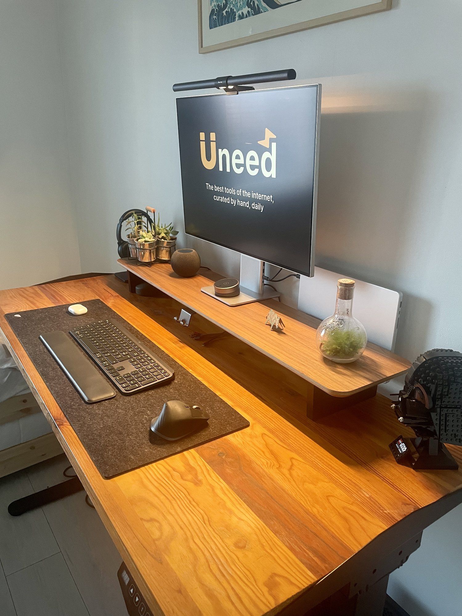 A modern and minimalist workspace featuring a wooden standing desk with a sleek ultra-wide monitor, small potted plants, and various neatly organised stationery items