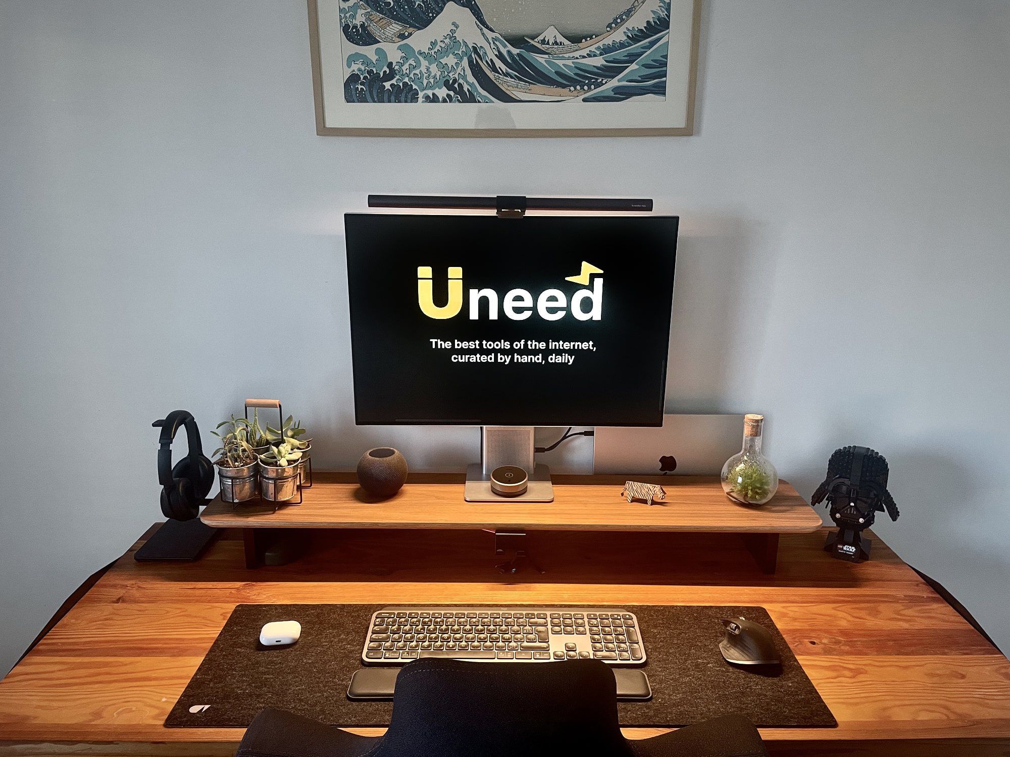 A small, cosy and inviting home office with a large wooden sit-stand desk that holds a monitor, laptop, keyboard, mouse, and a few decorative items, such as potted plants and a LEGO Star Wars Dark Trooper Helmet