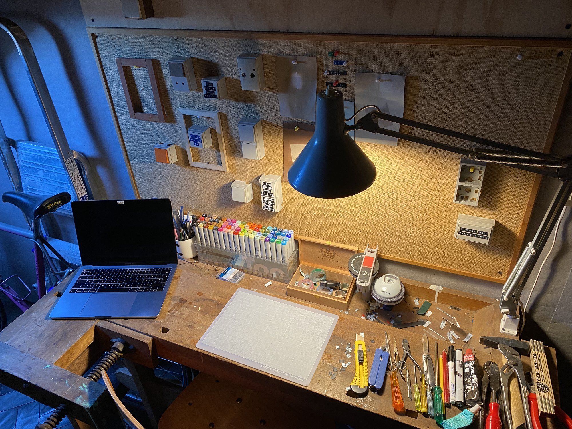 An artistic desk setup in the attic featuring a laptop, a lamp, a set of Conic pens, and other tools