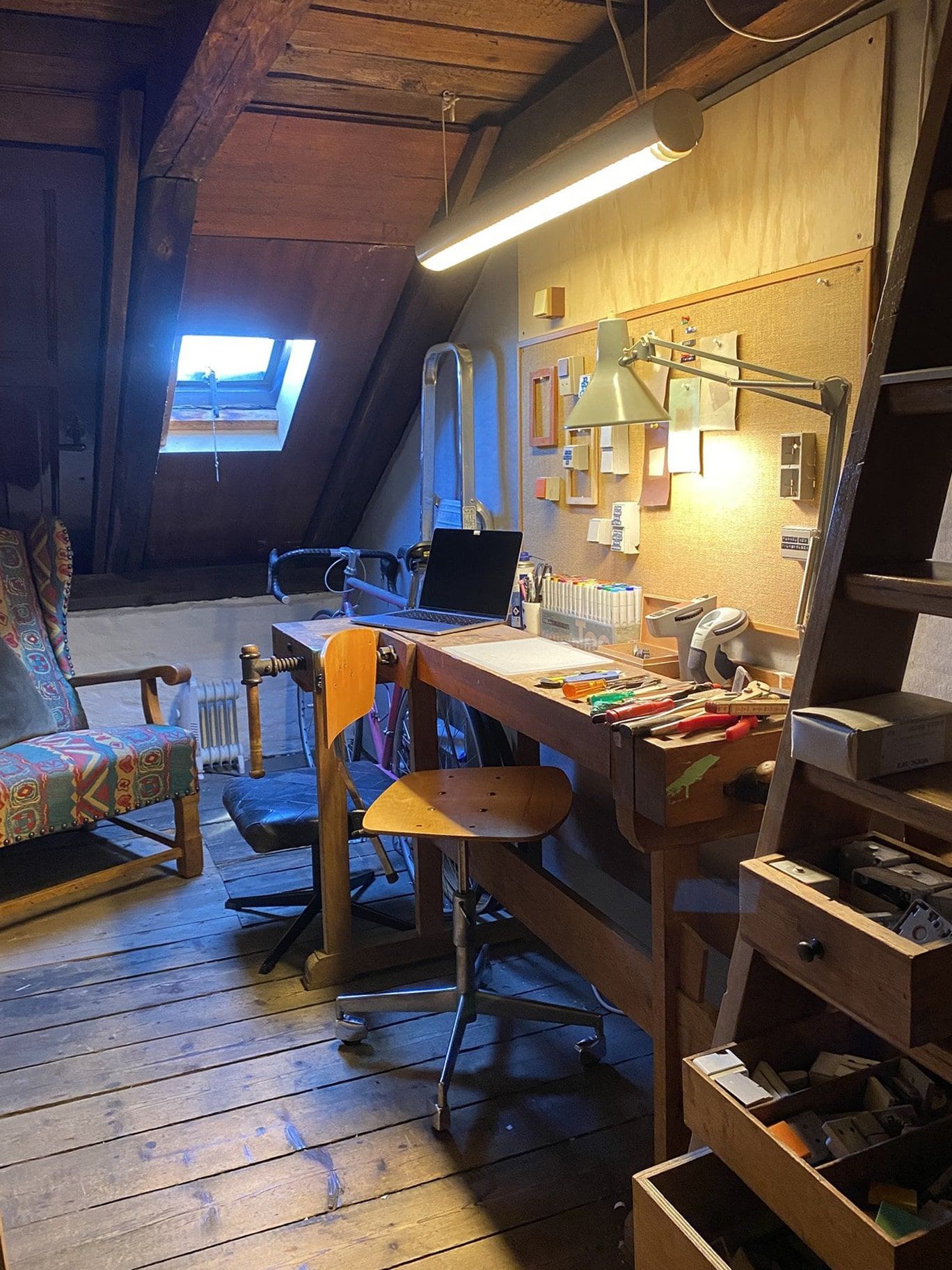 A rustic attic workspace with a laptop and a lot of artistic tools on a wooden desk. There is also a lamp on the desk and a skylight window in the roof