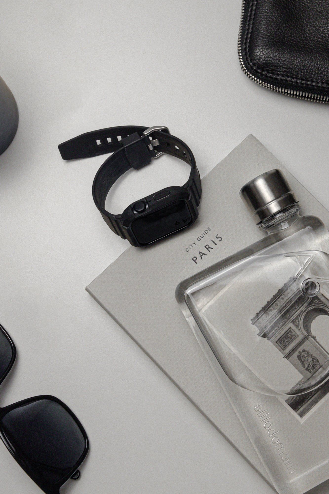A Paris city guide, sunglasses, a watch, and a transparent reusable water bottle arranged neatly on a white desk