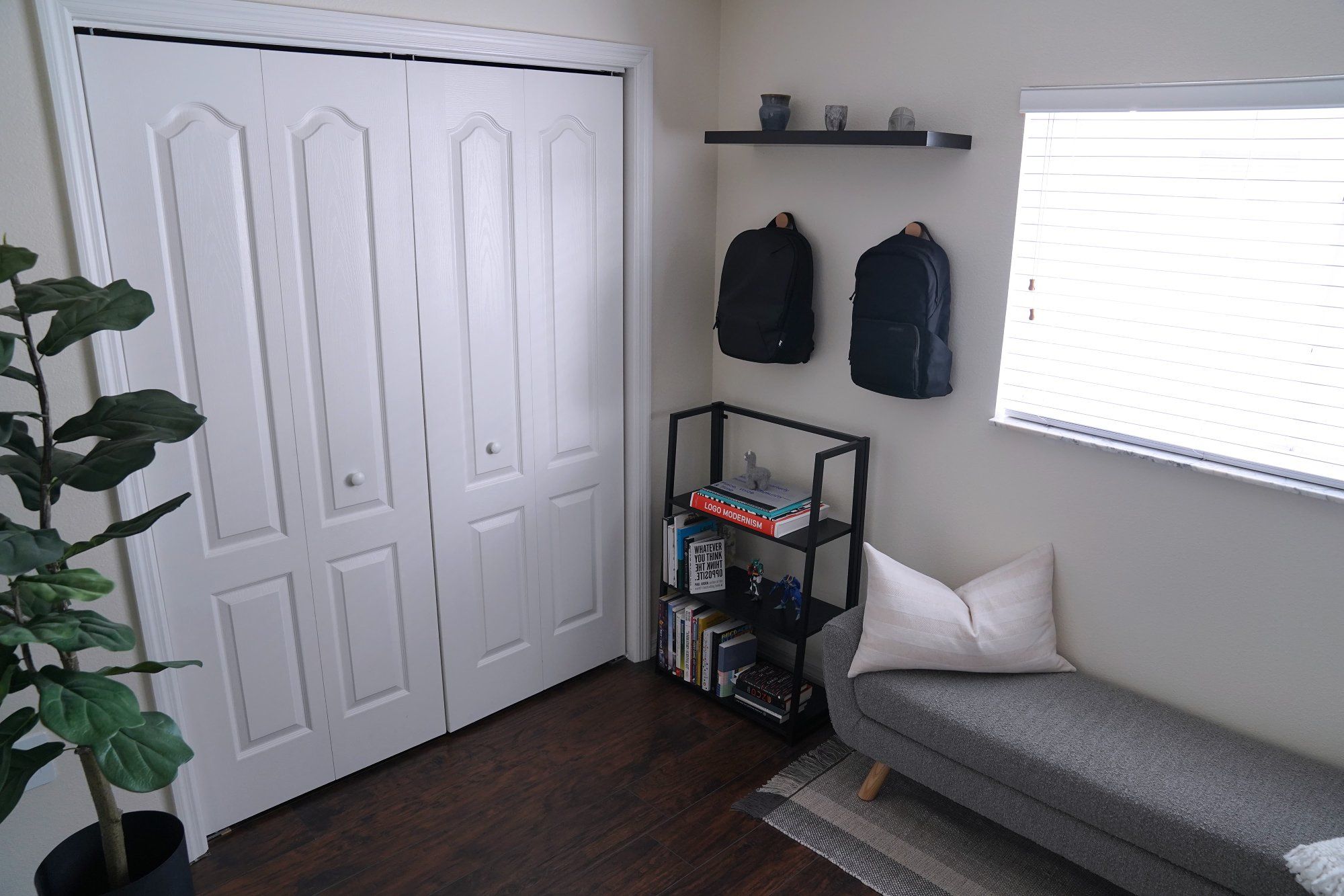 A minimal home office’s corner with a seating bench, a free-standing bookcase, two backpacks hanging on the wall, and an indoor plant