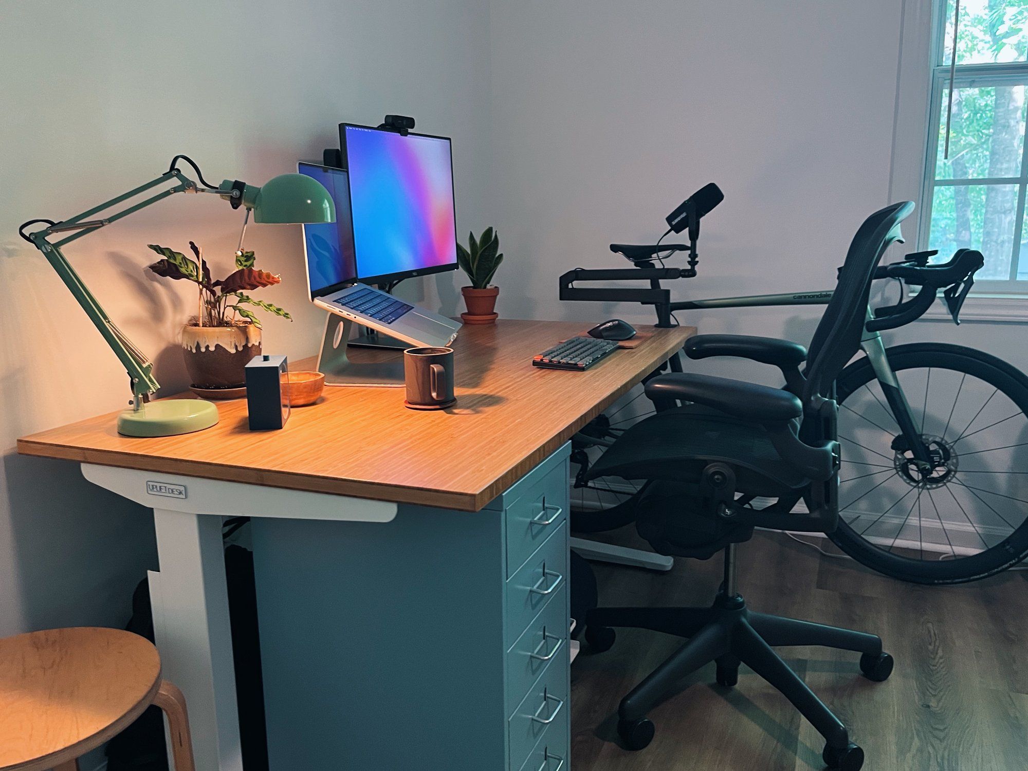 An view of a standing desk setup shows grey drawers, as well as an open MacBook Pro, an monitor, an olive table lamp, a mug, and some houseplants sitting on the desktop. Positioned between the desk and the window is a road bike