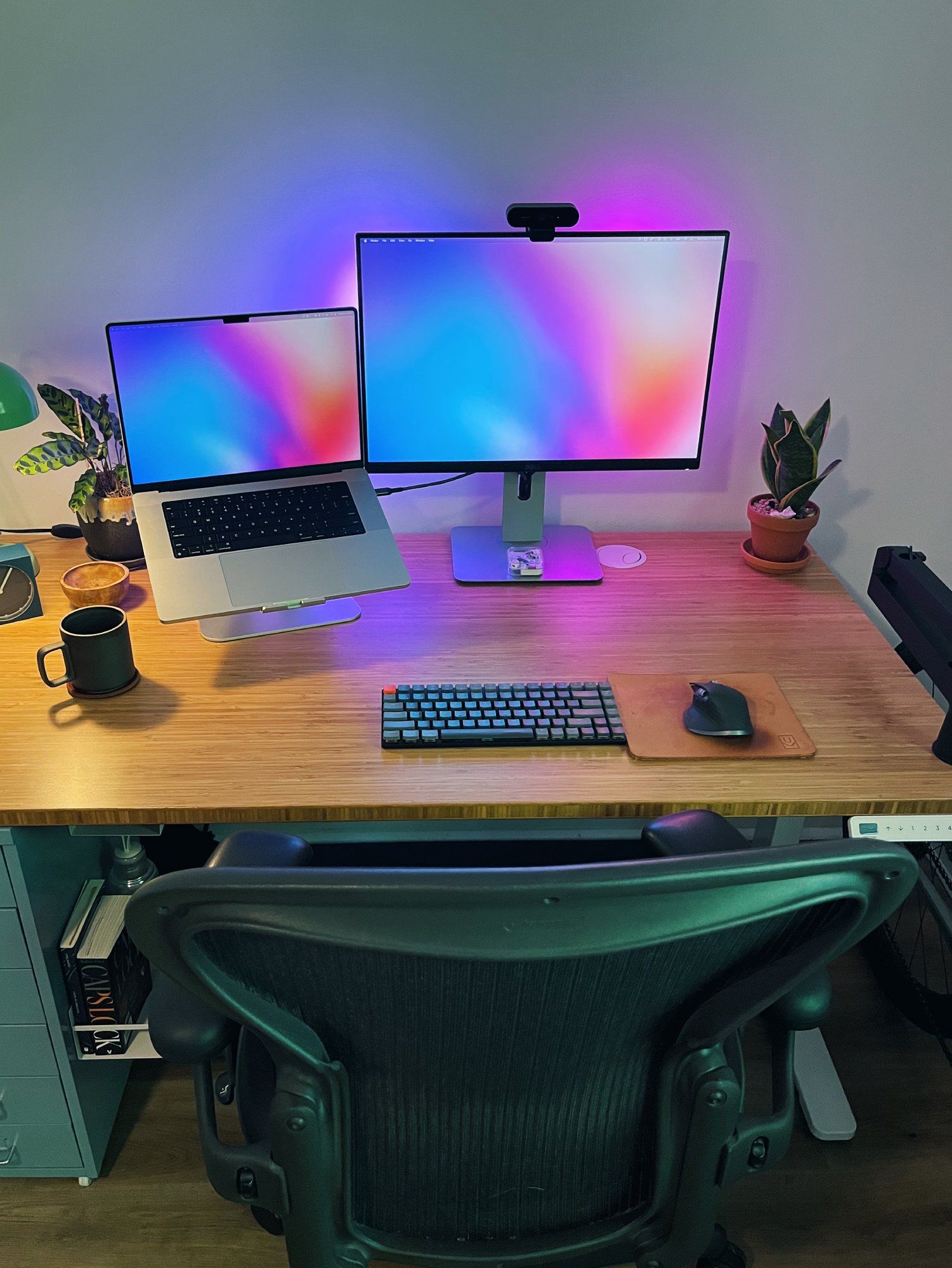 A minimal desk setup featuring a Dell UltraSharp monitor, a MacBook Pro laptop, a Keychron K3 keyboard, a Logitech MX3 mouse, and two houseplants