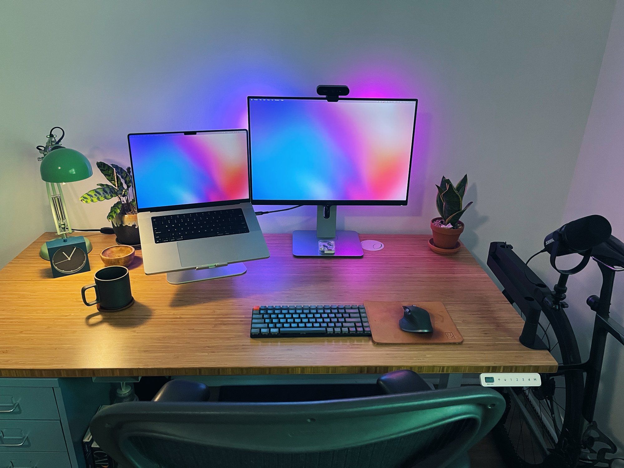A top view of a tidy designer workspace with a monitor, laptop, mechanical keyboard, mouse, desk lamp, and two houseplants on the desk