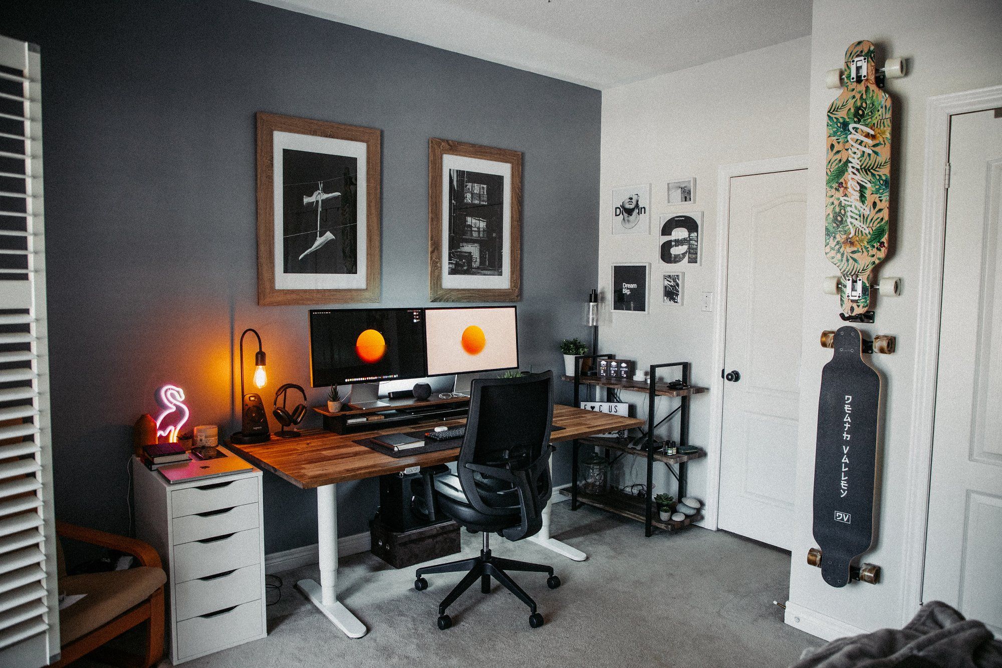 A standing desk setup featuring two Apple Studio Displays, a mechanical keyboard, a Lacasse office chair, and an ALEX drawer, along with several framed art posters and skateboards hanging on the walls