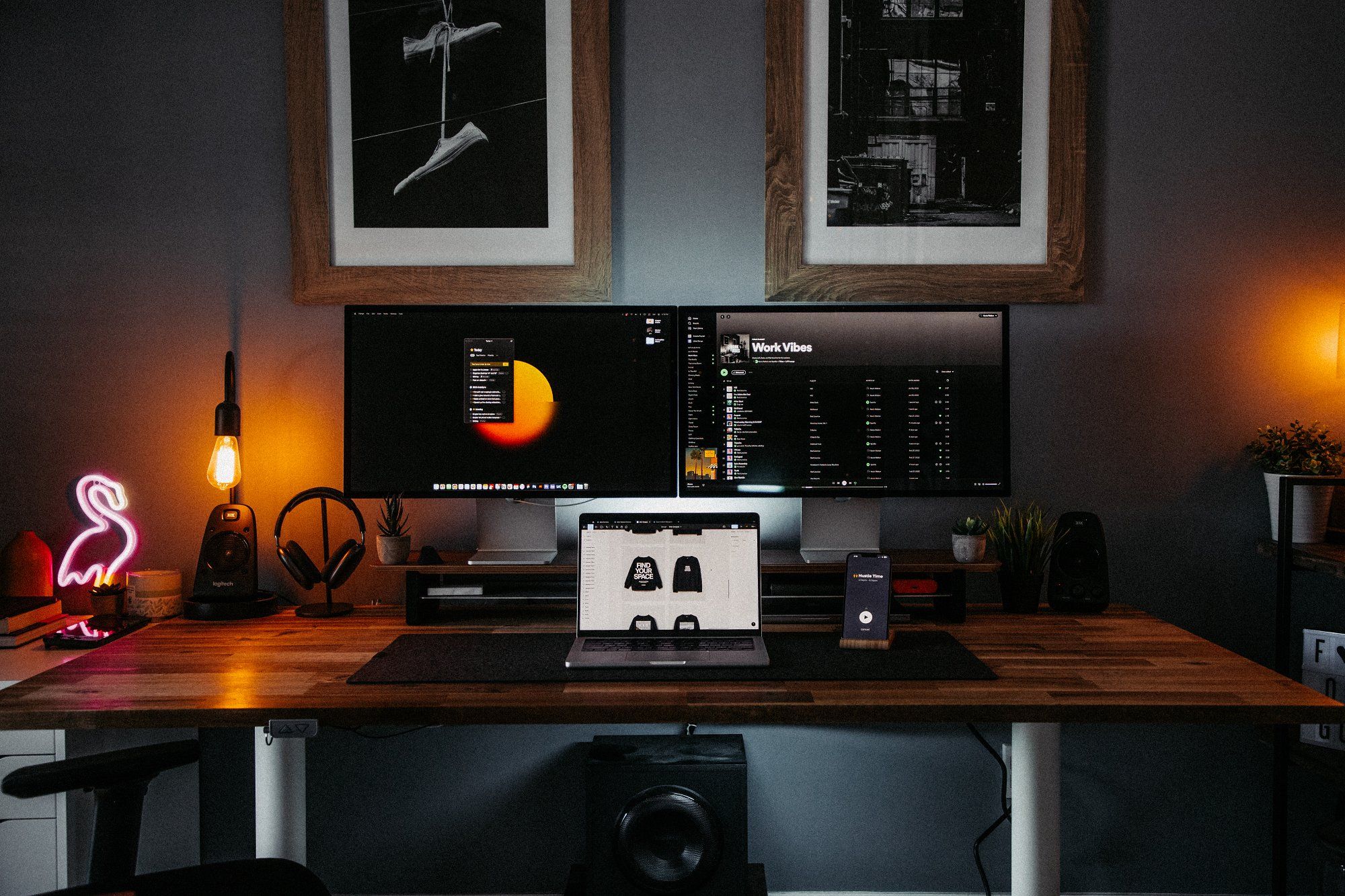 A home office setup with a MacBook, an iPhone, and two Apple Studio Displays on the desk, and two framed photo posters hanging on the wall above it