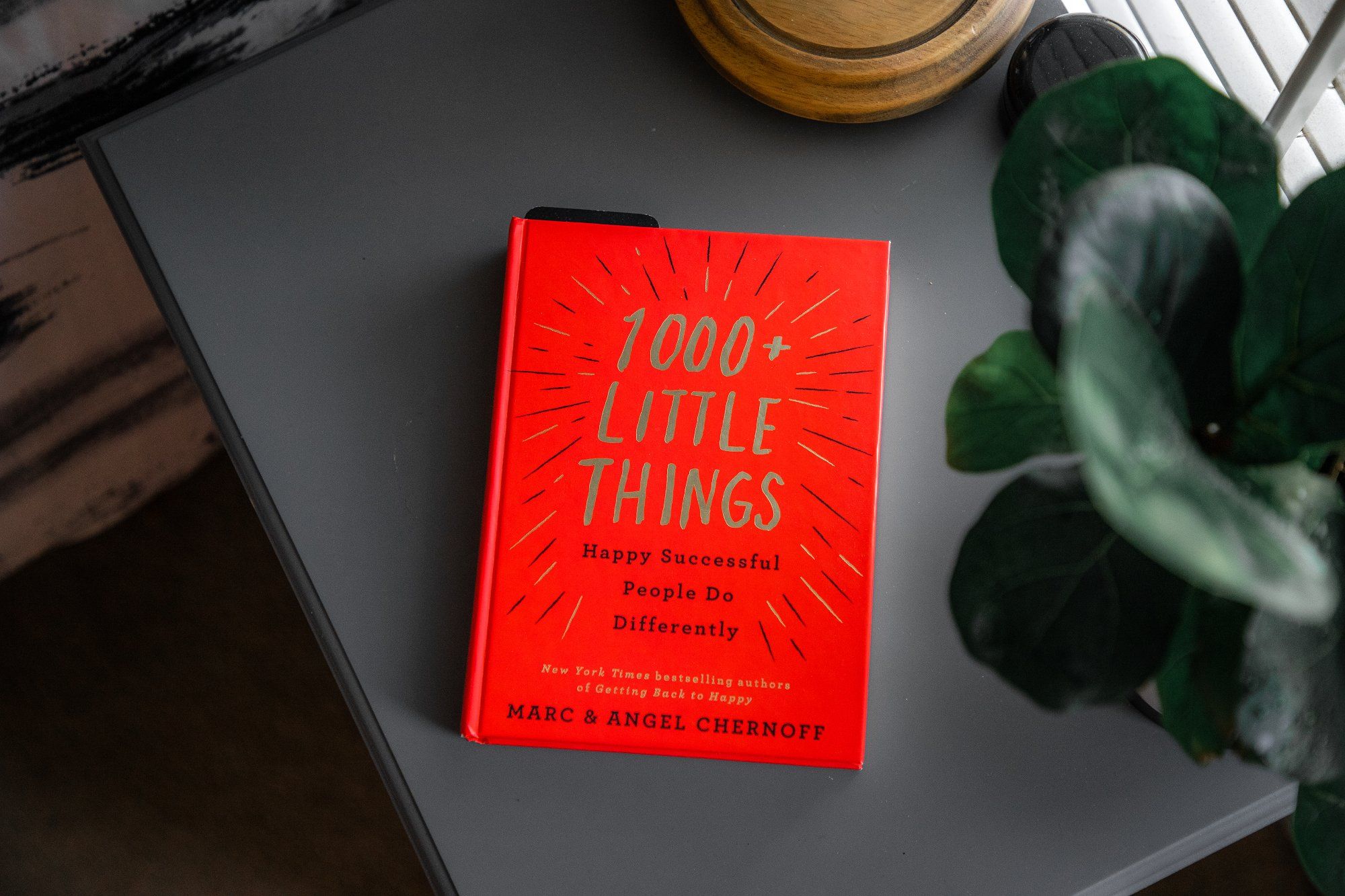 A 1000+ Little Things Happy Successful People Do Differently book by Marc & Angel Chernoff lying on a bookstand by the window