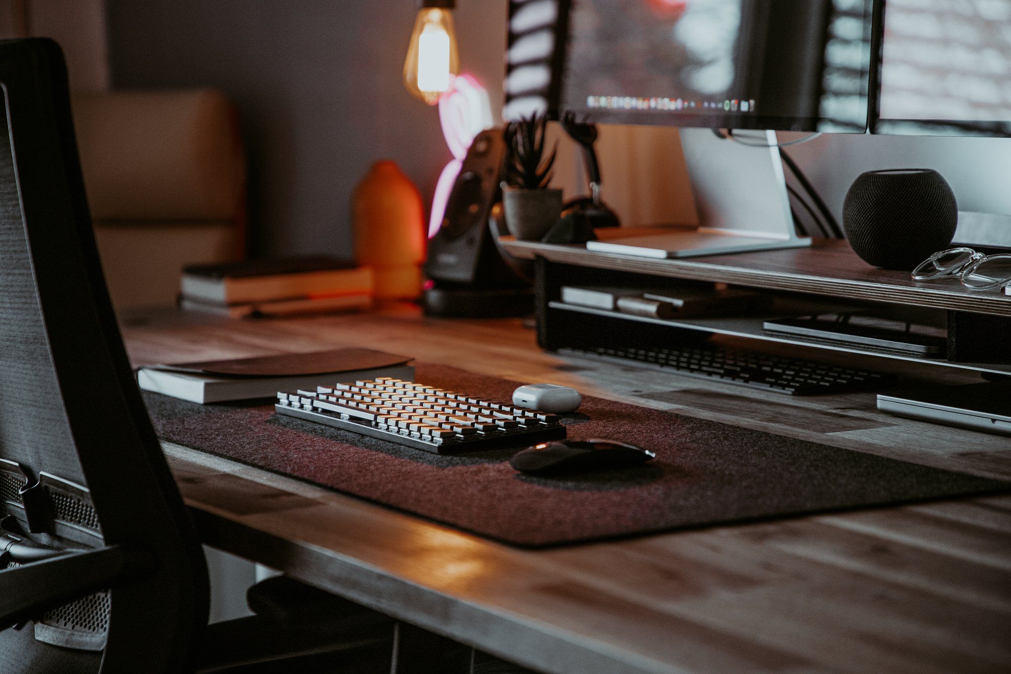 A mechanical keyboard, an Apple Magic Mouse, AirPods in a case, and a notebook are placed on a Grovemade desk mat in a neat desk setup in darker tones