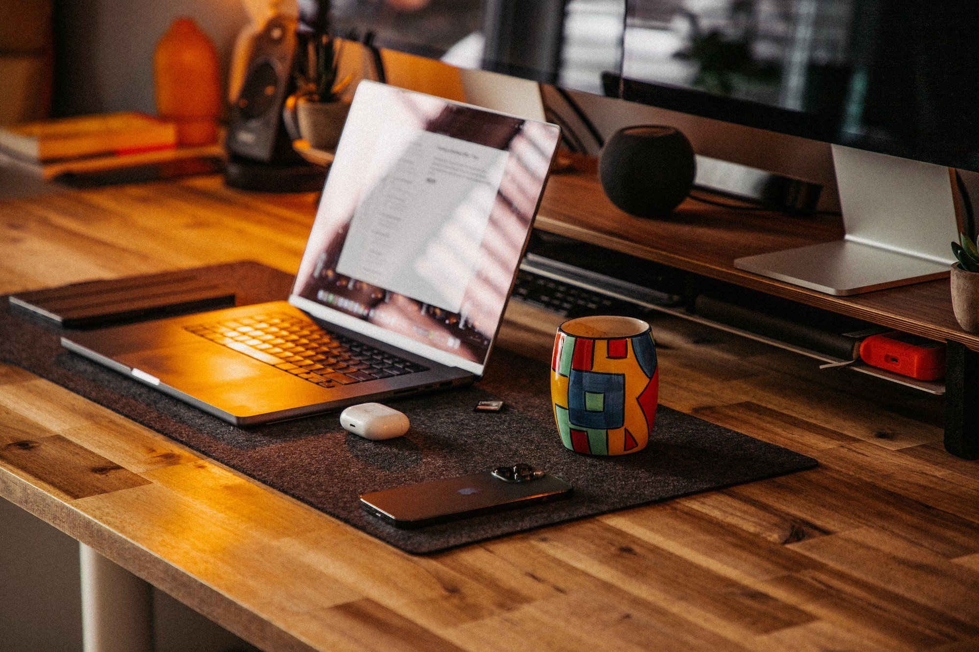 On a desk with a wooden top lies a wool Grovemade desk mat, and on it are placed a MacBook Pro, an iPhone, AirPods, and a mug with a multicoloured pattern