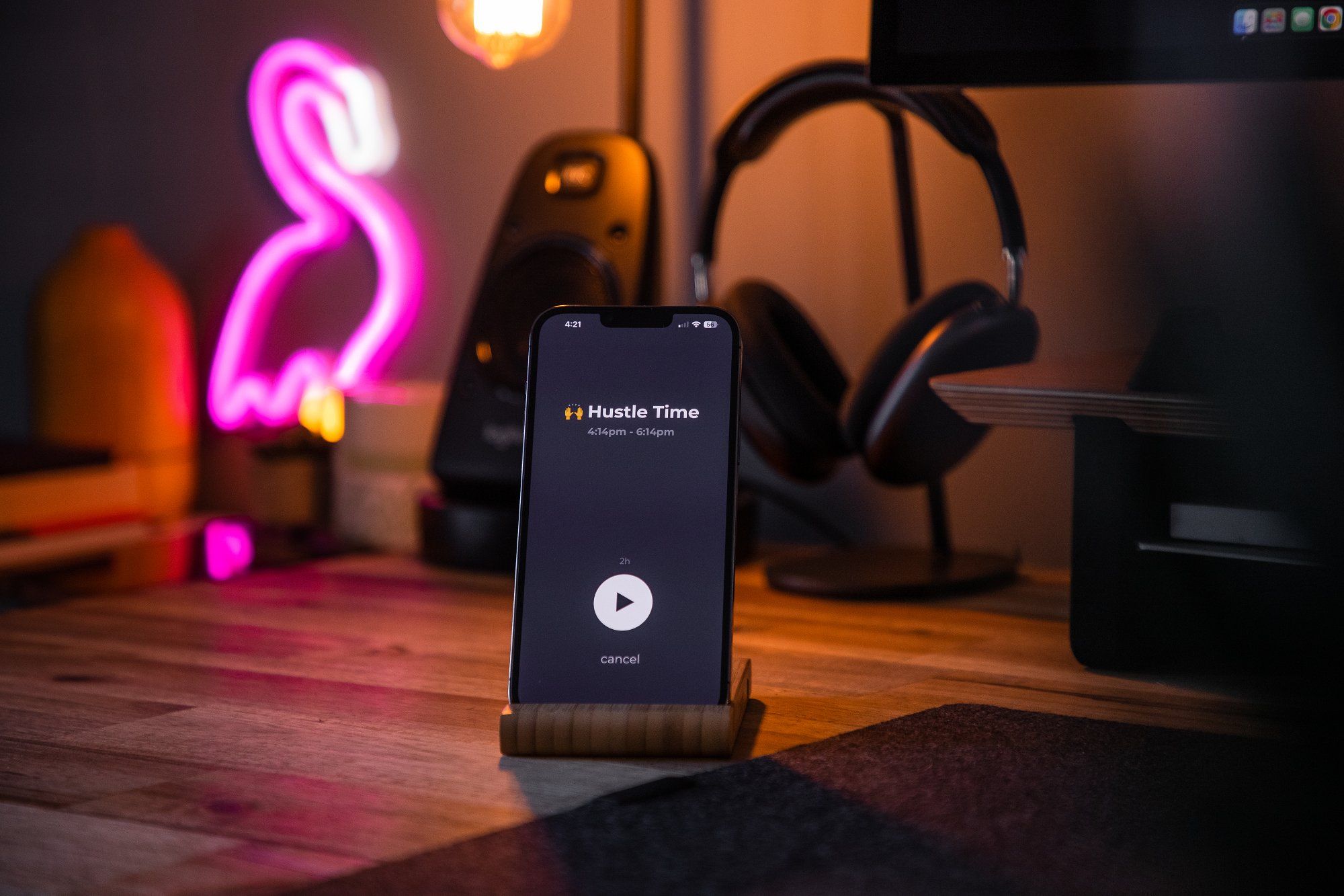 An iPhone is mounted on a stand with a hustle timer on the screen, and an LED flamingo sign along with headphones are behind it