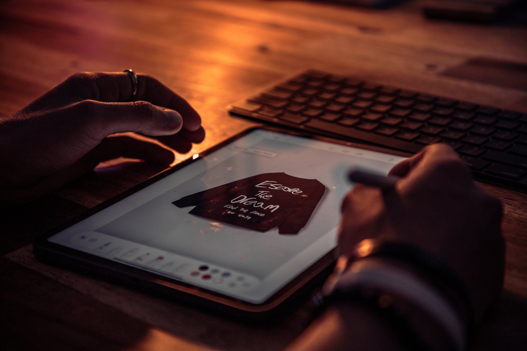 Under the light of a lamp, the hands of a man are shown creating a sweater design on the iPad Mini screen. A mechanical keyboard can be seen in the background