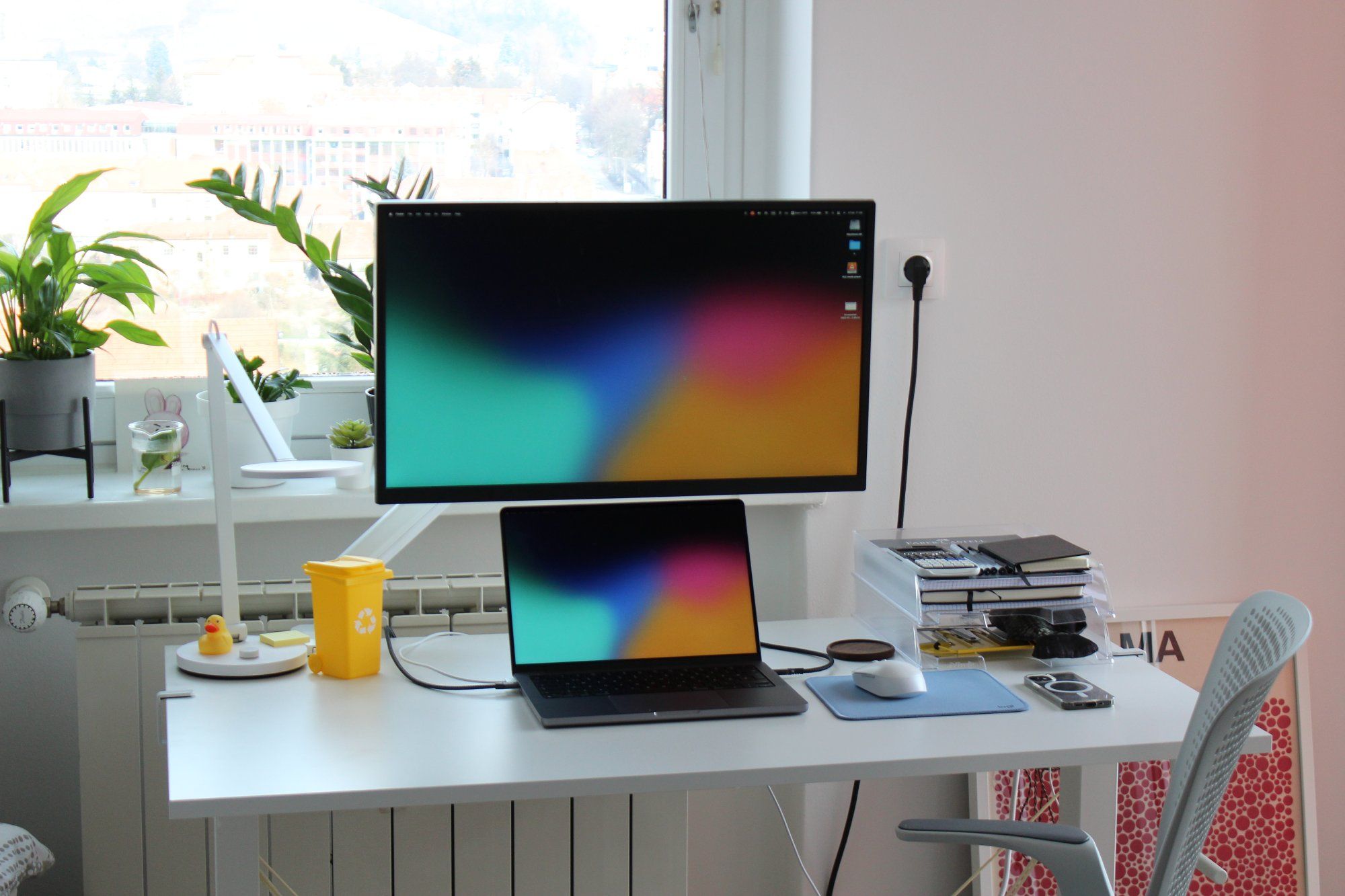 A home office setup featuring an IKEA TROTTEN sit-stand desk, a 4K LG monitor, an Apple MacBook Pro, a desk lamp, a mouse on the mat, books, notepads, and other accessories and peripherals