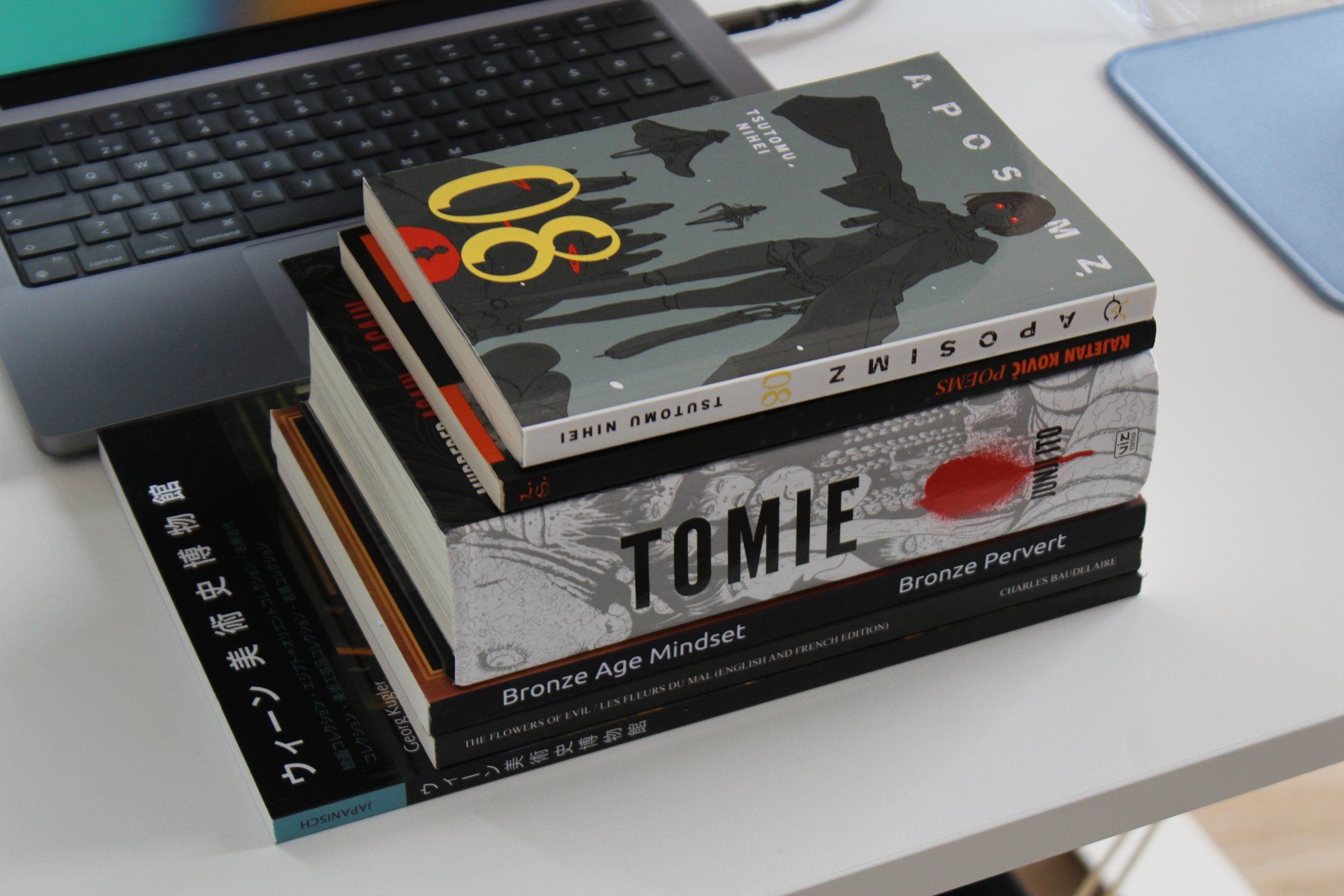 A stack of books, including mangas Tomie by Junji Ito and Aposimz by Tsutomu Nihei, as well as poetry collections by Baudelaire and Kajetan Kovič, lie on the desk