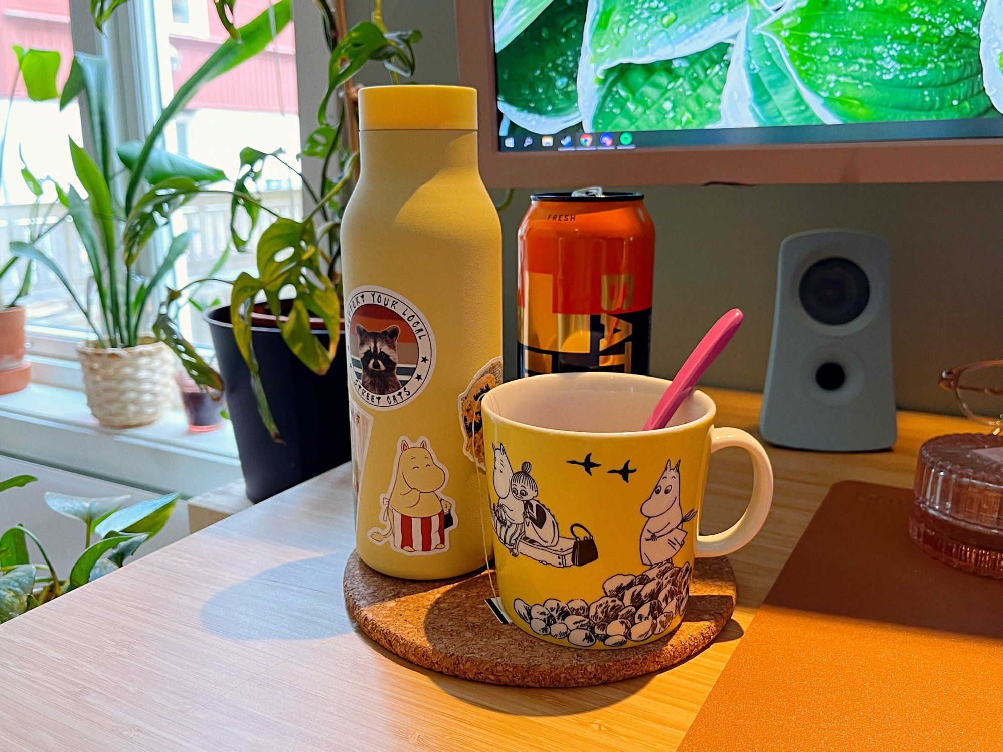 A yellow Moomin water bottle, a can of Battery energy drink, and a yellow Moomin mug on a cork table mat