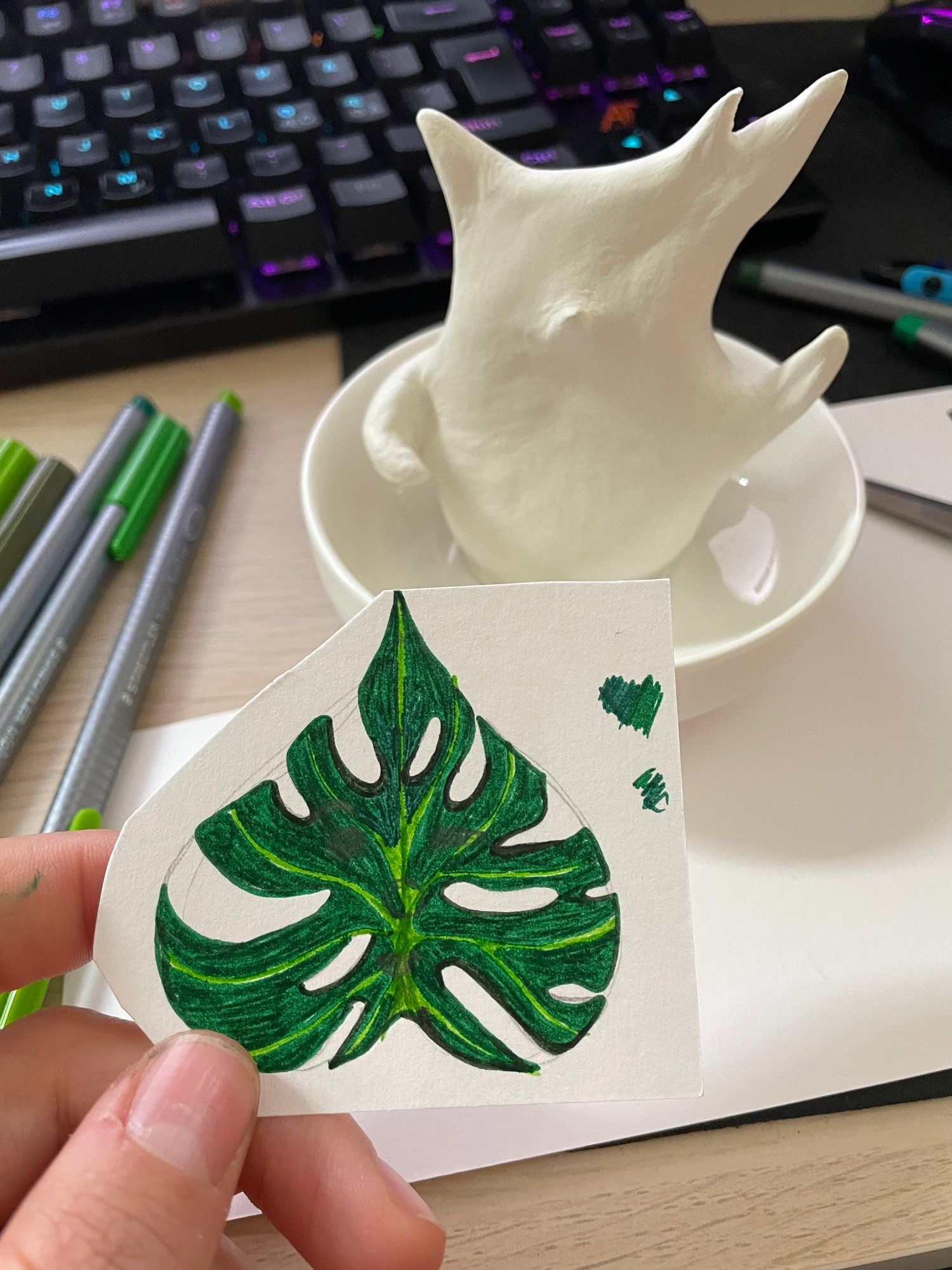 A monstera leaf drawn on a piece of paper in the foreground, and a white figurine on a saucer in the background