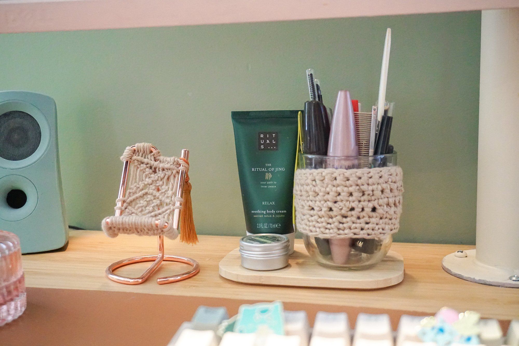 A phone stand with a handmade macramé cradle, a makeup glass with a crocheted band, a tube of Rituals soothing body cream, a lip balm, and a teal hand-painted Logitech speaker on the desk in a home office