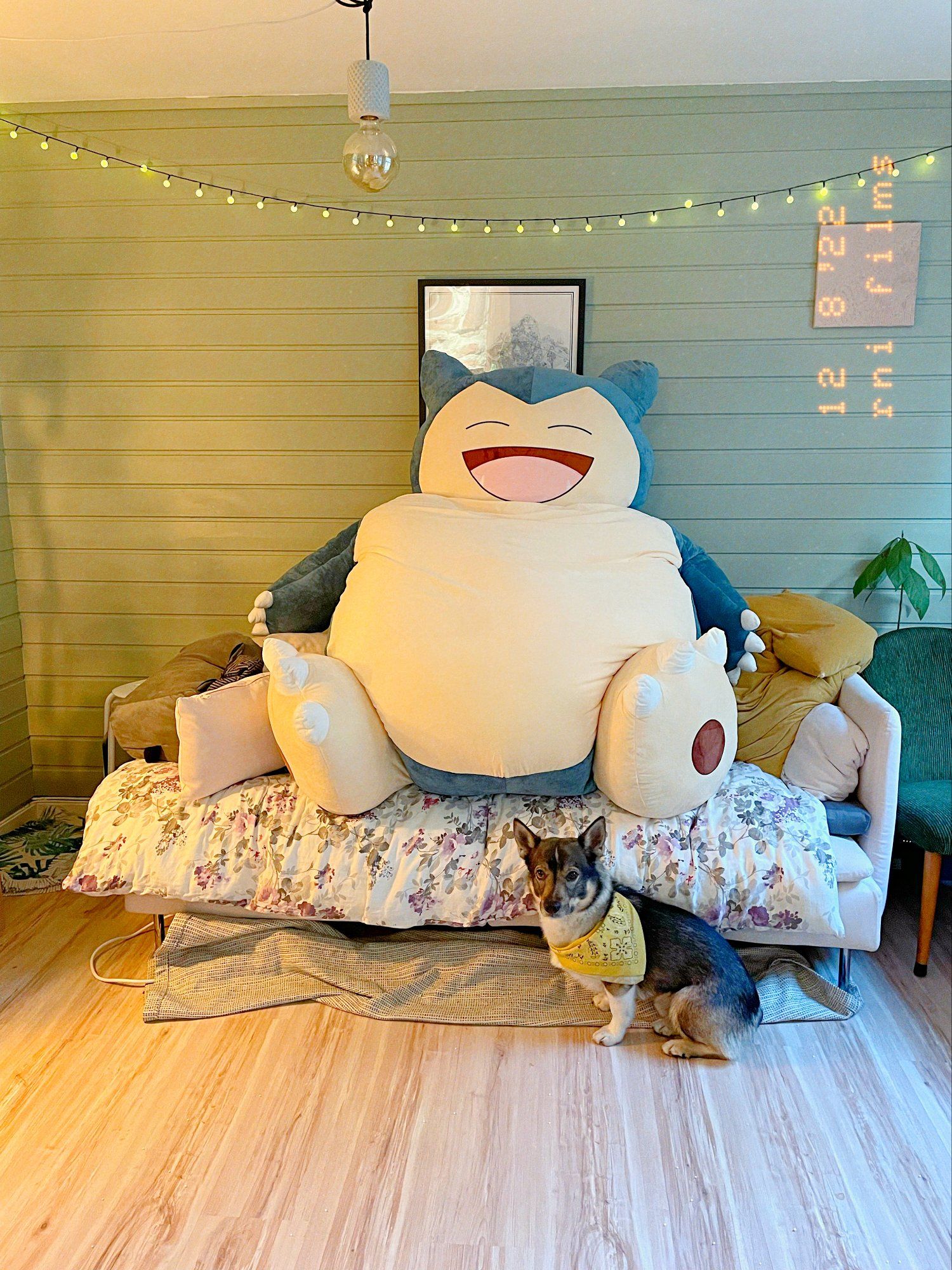 A photo with a dog in a yellow neckerchief sitting on the floor, and a huge Snorlax plush toy Pokèmon sitting on the couch