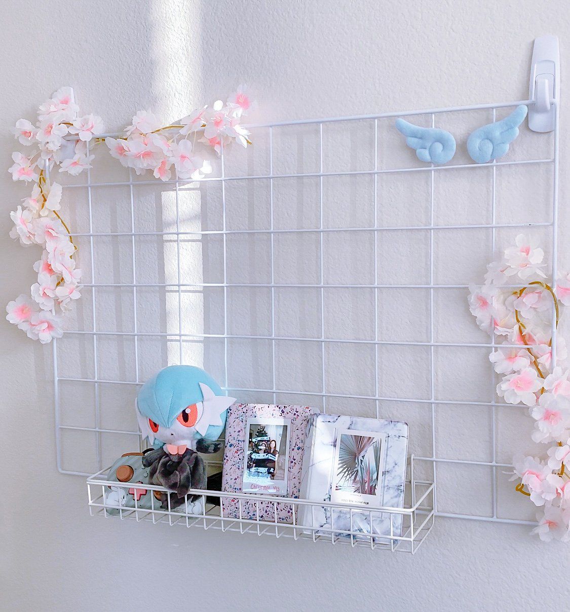 A white wall grid with artificial Sakura flowers, photos, a Shiny Mega Gardevoir Pokémon plush toy, and a game console joystick 