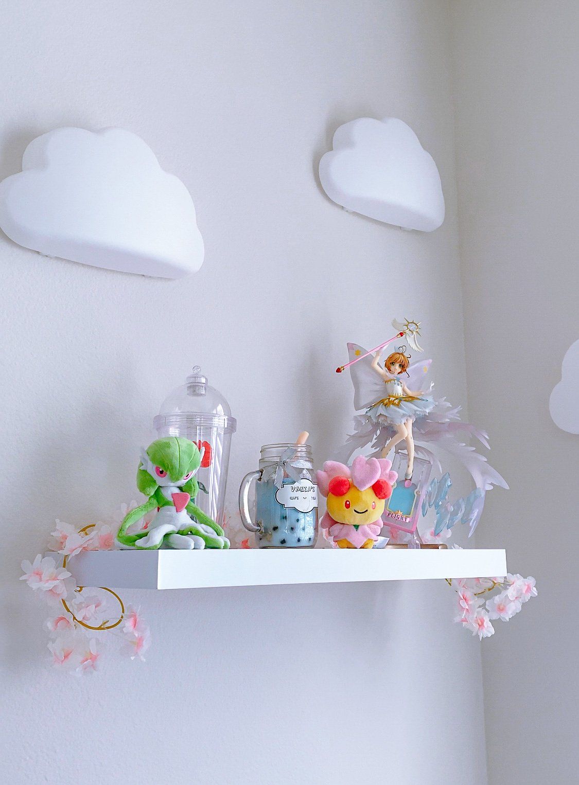 Gardevoir and Cherrim Pokémon plush toys, a Sakura Kinomoto action figure, and a jar candle along with some artificial Sakura flowers on a floating shelf