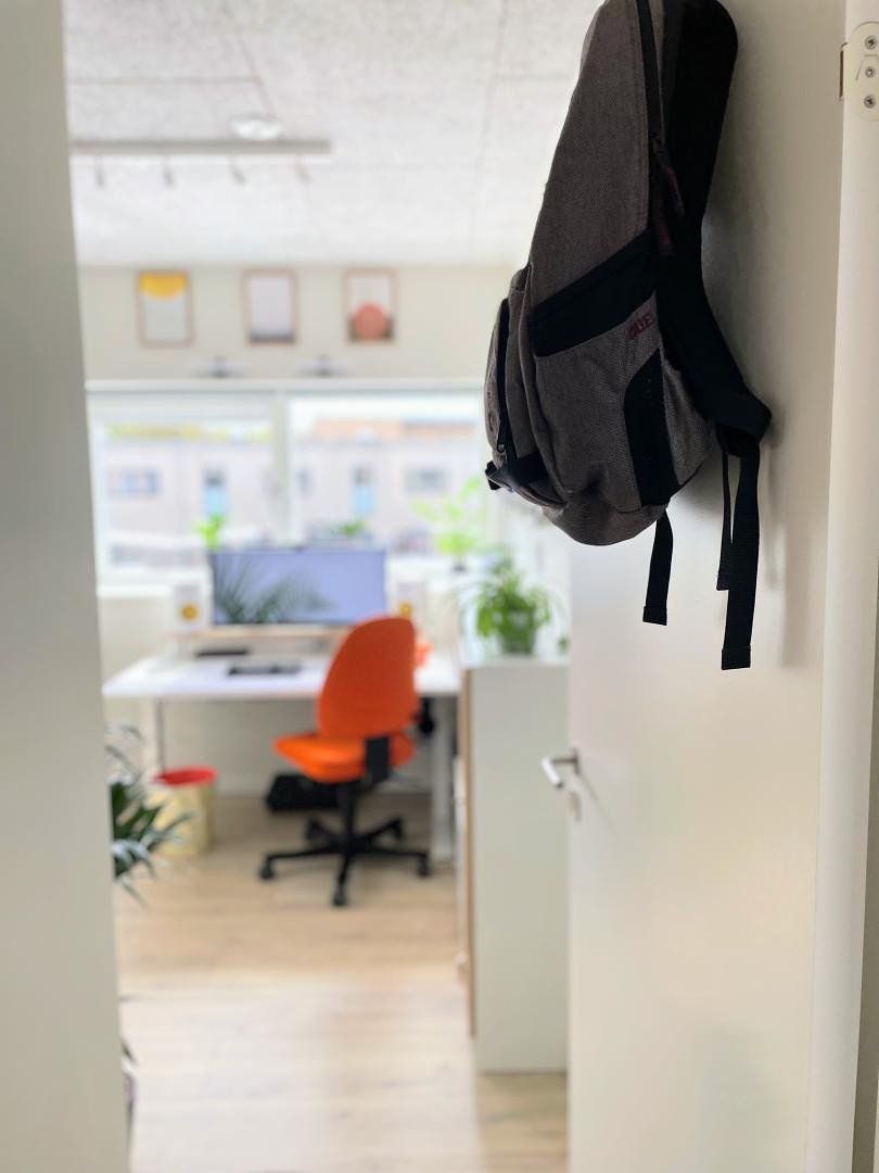 In the foreground of the photo, there is an open door with a backpack hanging on it; in the background, a bright desk setup is faintly visible in the bokeh