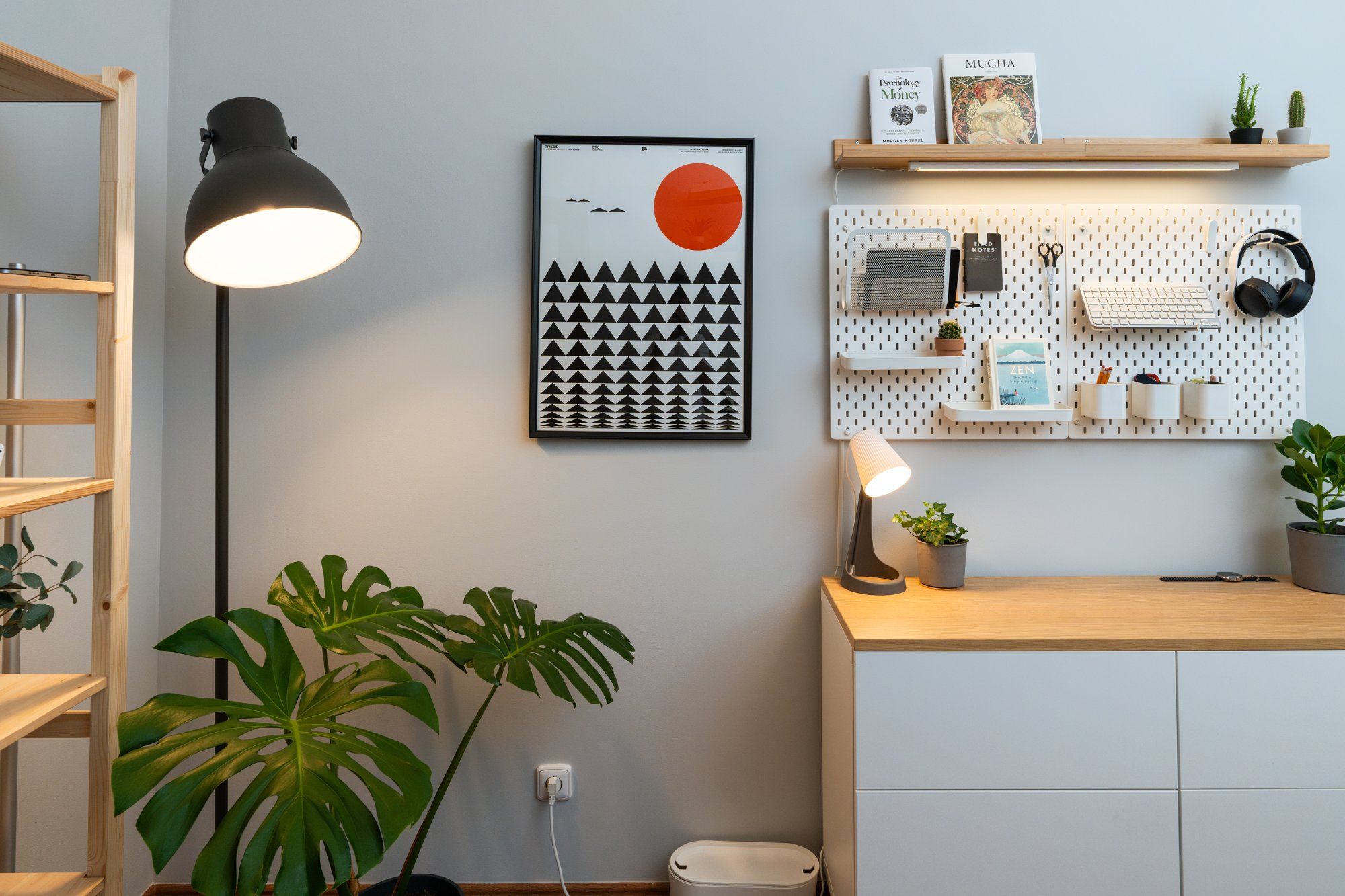 A home office corner with a wooden bookcase, an IKEA floor lamp, a large Monstera, and two pegboards on the wall