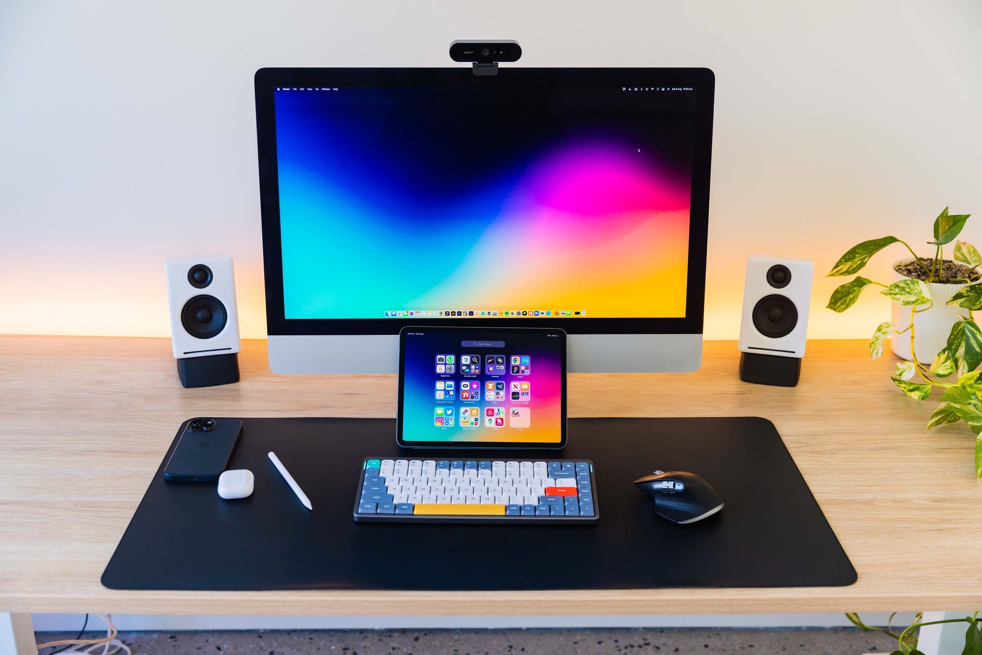 An iMac, an iPad with an Apple Pencil, AirPods, an iPhone, a NuPhy Air75 keyboard, a Logitech MX Master 3 mouse, Audioengine A2+ speakers, and a potted houseplant on the desk