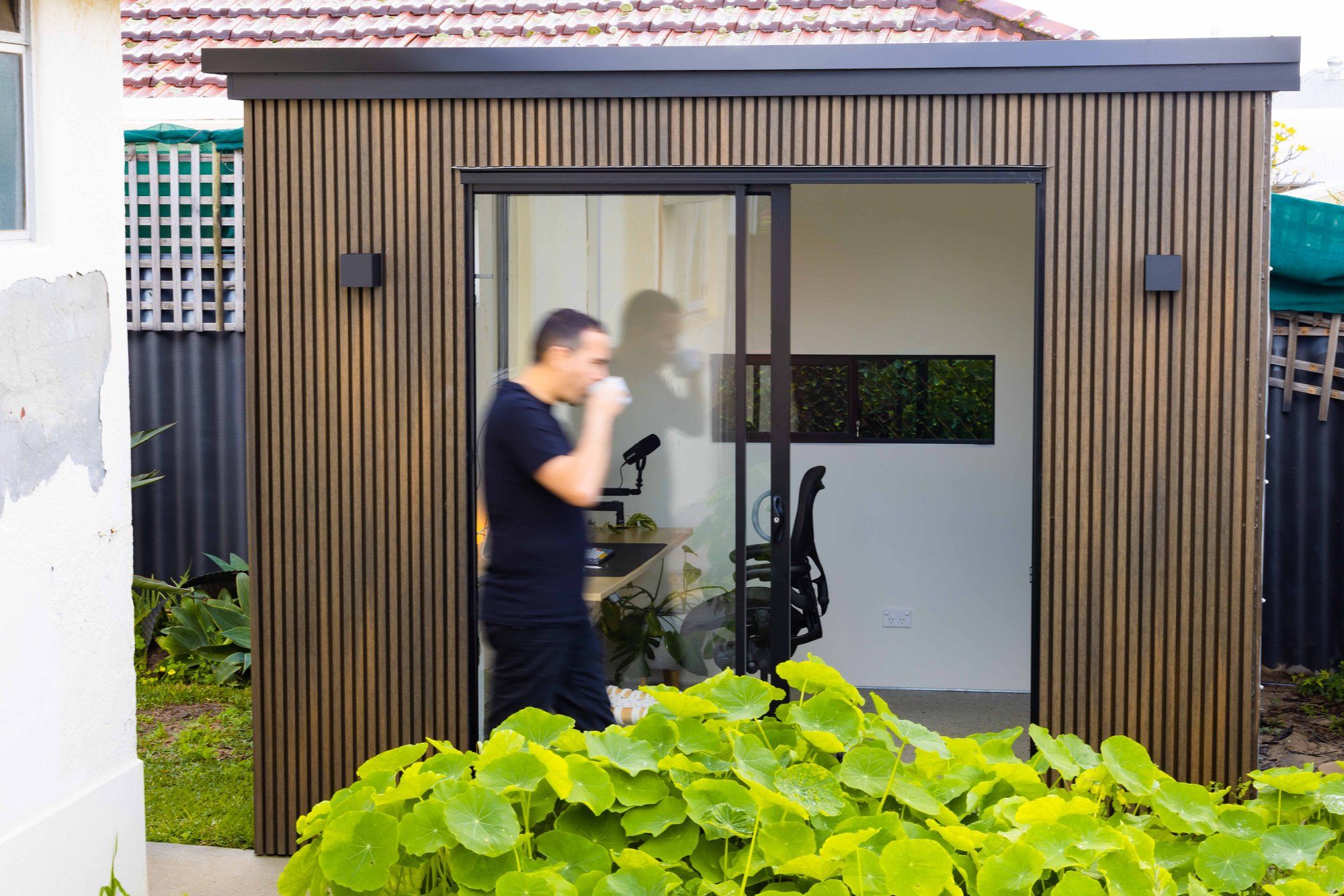 A man swiftly moving with a cup of coffee in hand, walking by his backyard office pod