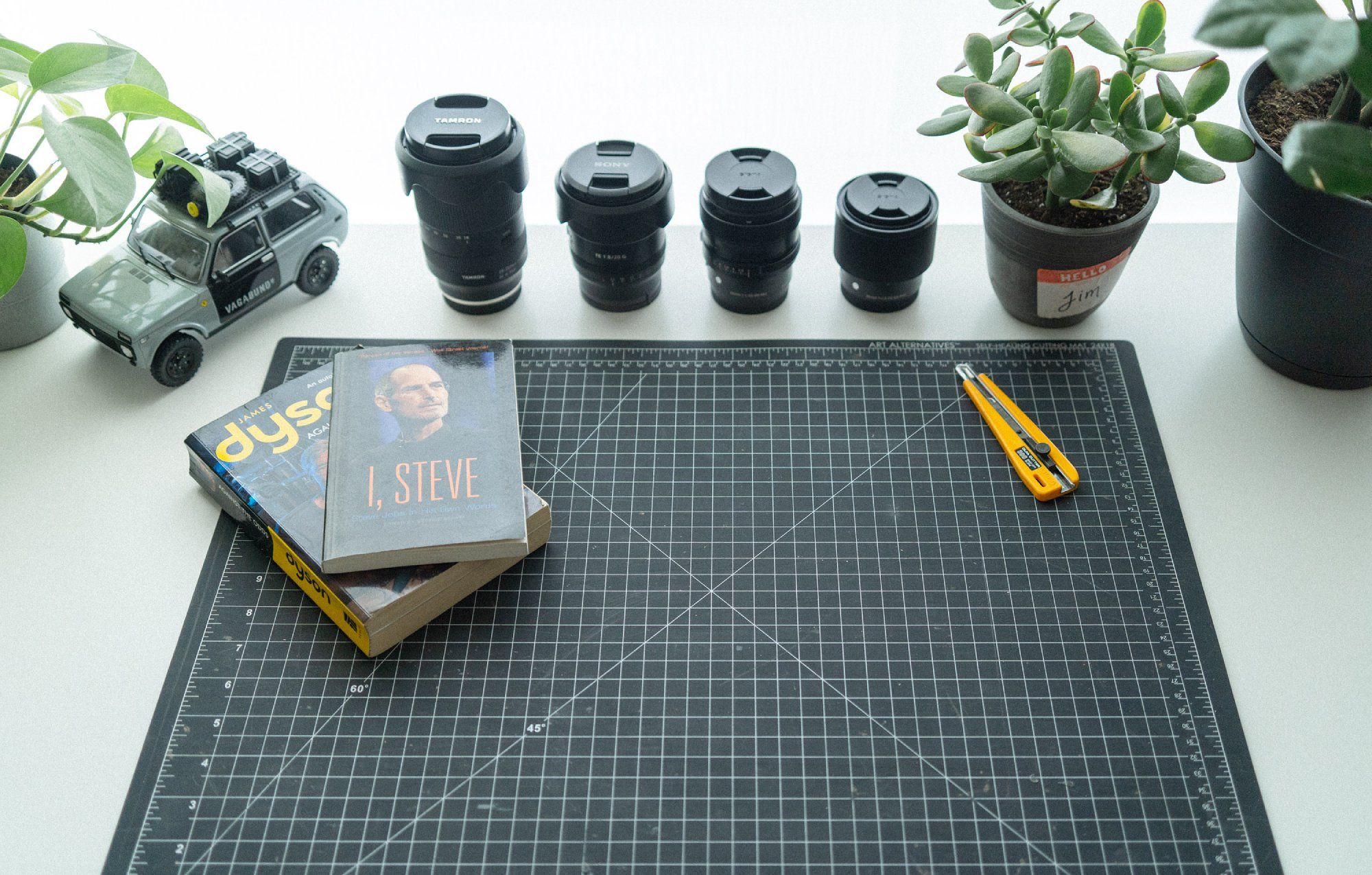 Two books and a ratchet-lock utility knife lying on a cutting mat next to 4 camera lenses, a model toy car, and some plants