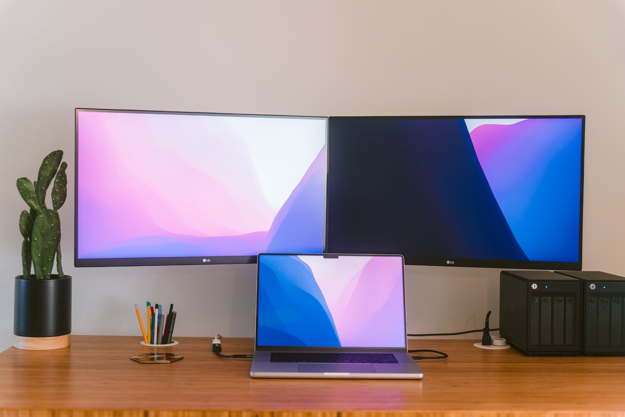 A close-up view of a sleek home office desk featuring dual LG monitors, an open laptop with gradient wallpaper, potted cactus plant, assorted pens, and computer accessories on a wooden surface