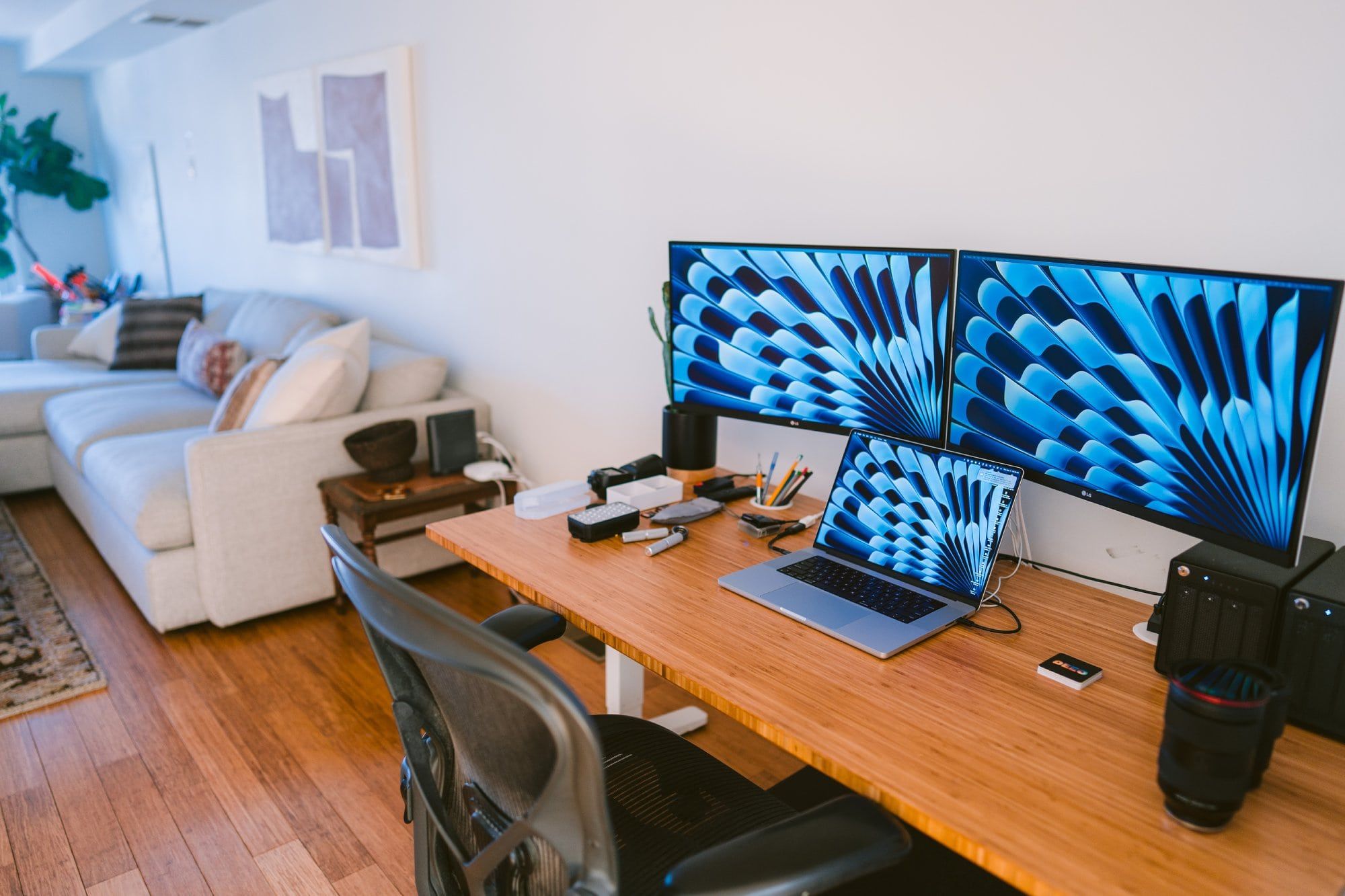 A contemporary home office setup featuring a dual-monitor display with vibrant blue patterns, alongside a laptop on a wooden desk, ergonomic chair, and cosy living room area in the background