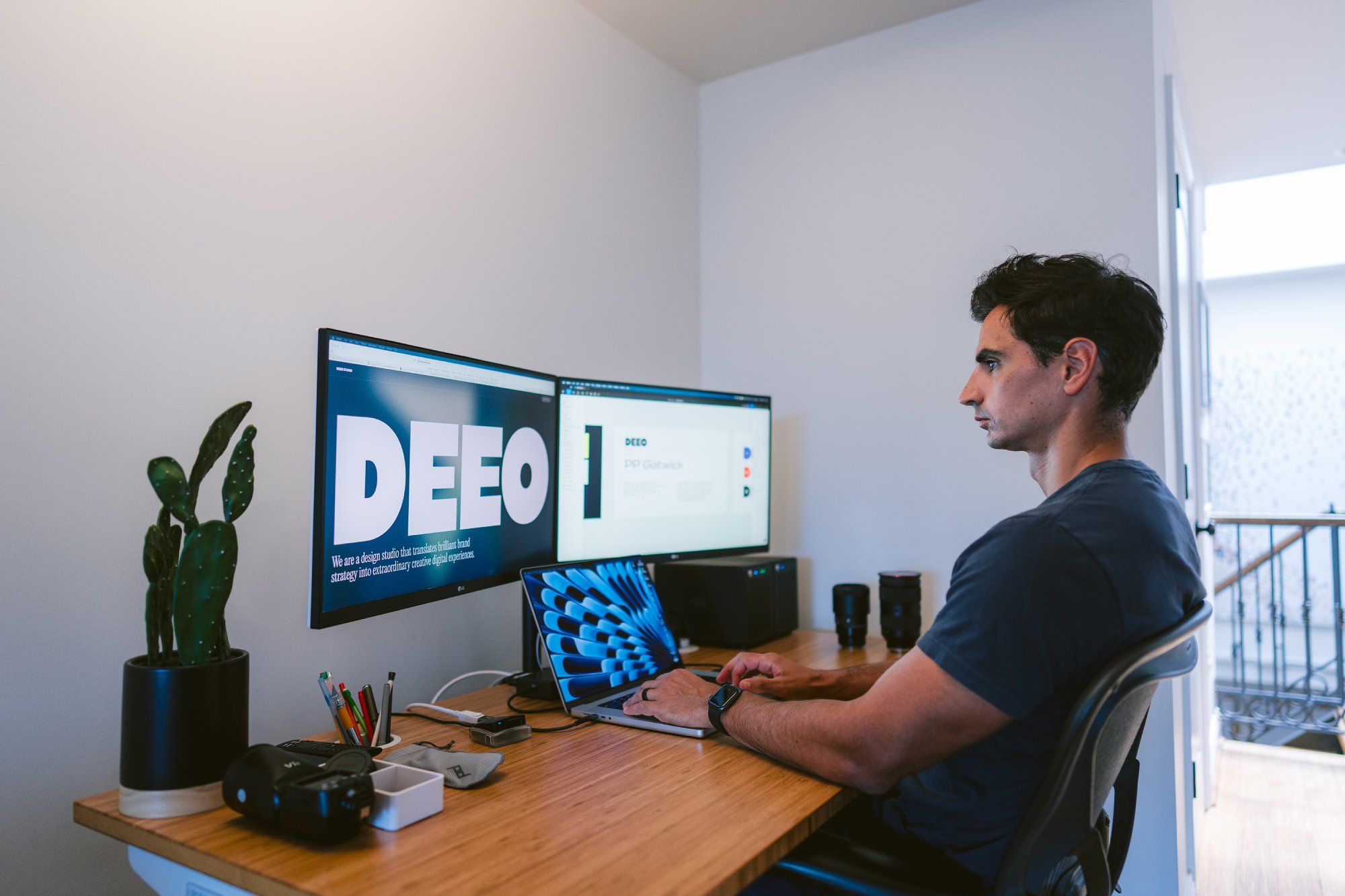 A man sitting at a desk with multiple computer monitors displaying graphics and text. The desk has various office items, including a laptop, a plant, and stationery. The room features a modern interior with a white wall background