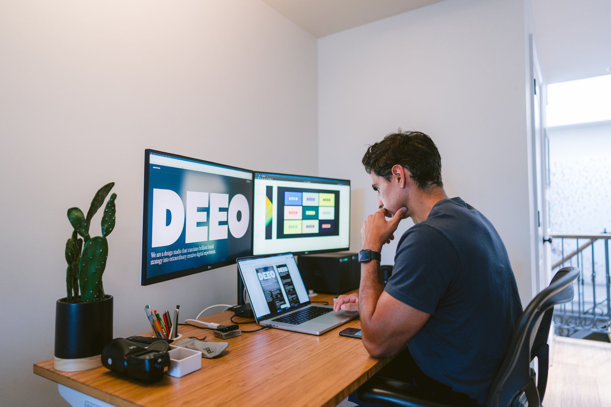A man is sitting at a wooden desk, deeply engrossed in his work. He is facing a set of dual monitors which display a prominent text DEEO and some design layouts