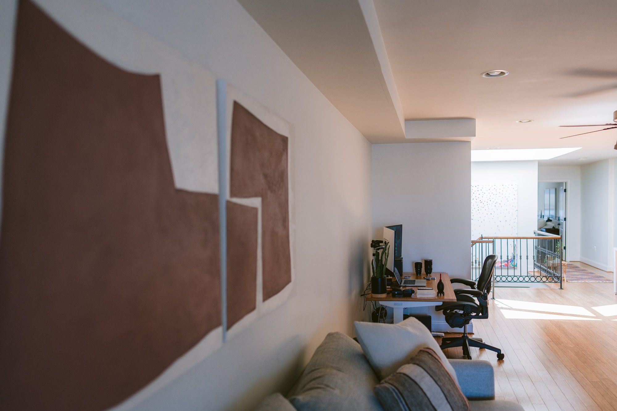 A spacious living area featuring an abstract brown and white wall art, with a view of a well-organised home office setup in the background, alongside a comfortable seating area and a balcony entrance with decorative railings