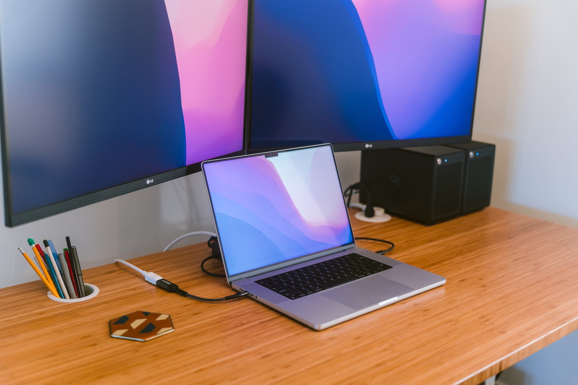 A contemporary workspace featuring two large LG monitors with gradient wallpapers, a sleek laptop with a similar wallpaper, an assortment of colored pens in a holder, a decorative coaster, and a couple of compact desktop speakers. The devices are neatly arranged on a wooden table