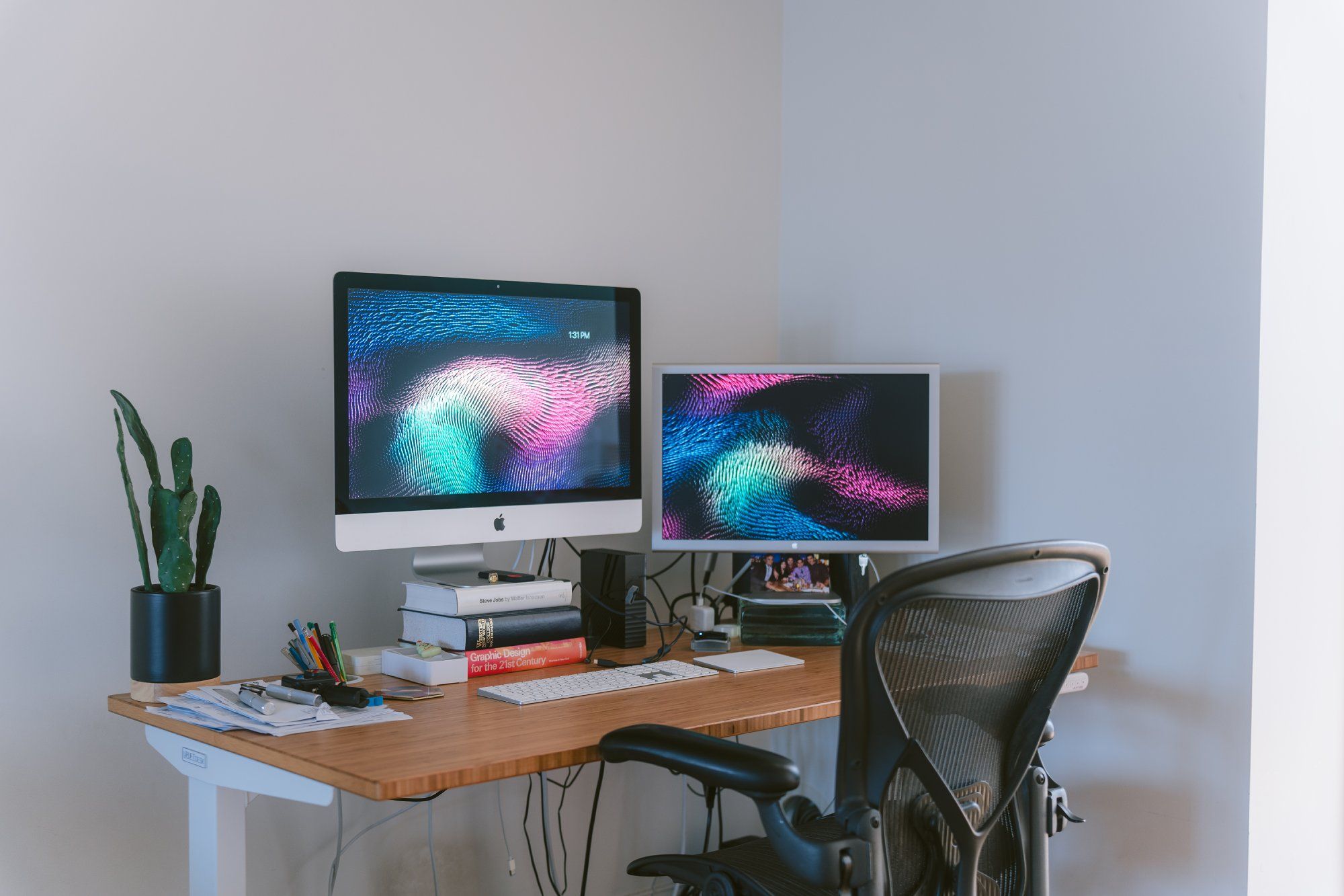 An organised home office setup showcasing an Apple iMac computer with a vibrant wave-patterned wallpaper, a secondary monitor displaying the same wallpaper, a potted cactus plant, a stack of design-related books, assorted writing utensils, and a comfortable ergonomic chair