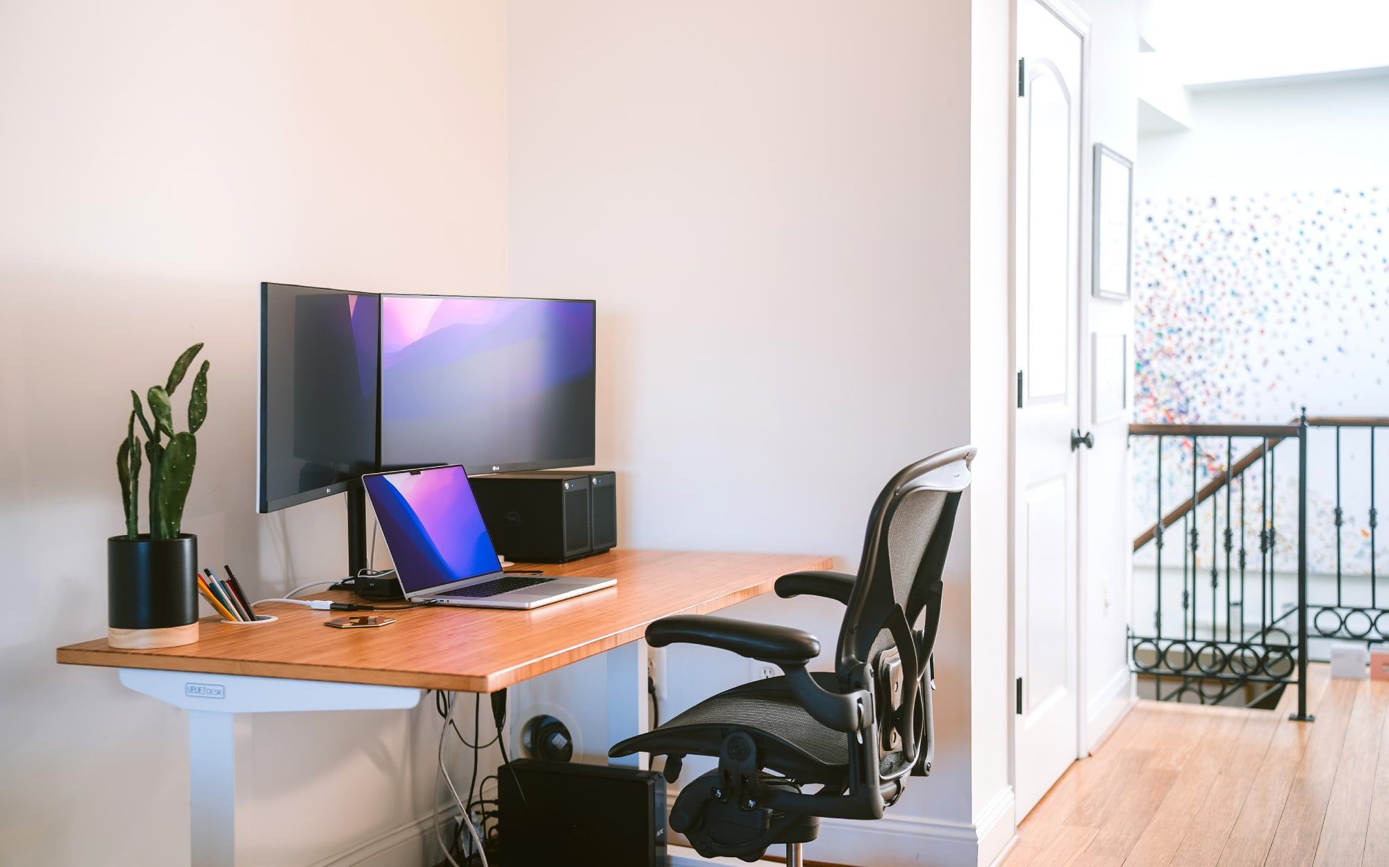 A modern home office setup with dual monitors, laptop on wooden desk, ergonomic chair, and a potted indoor plant