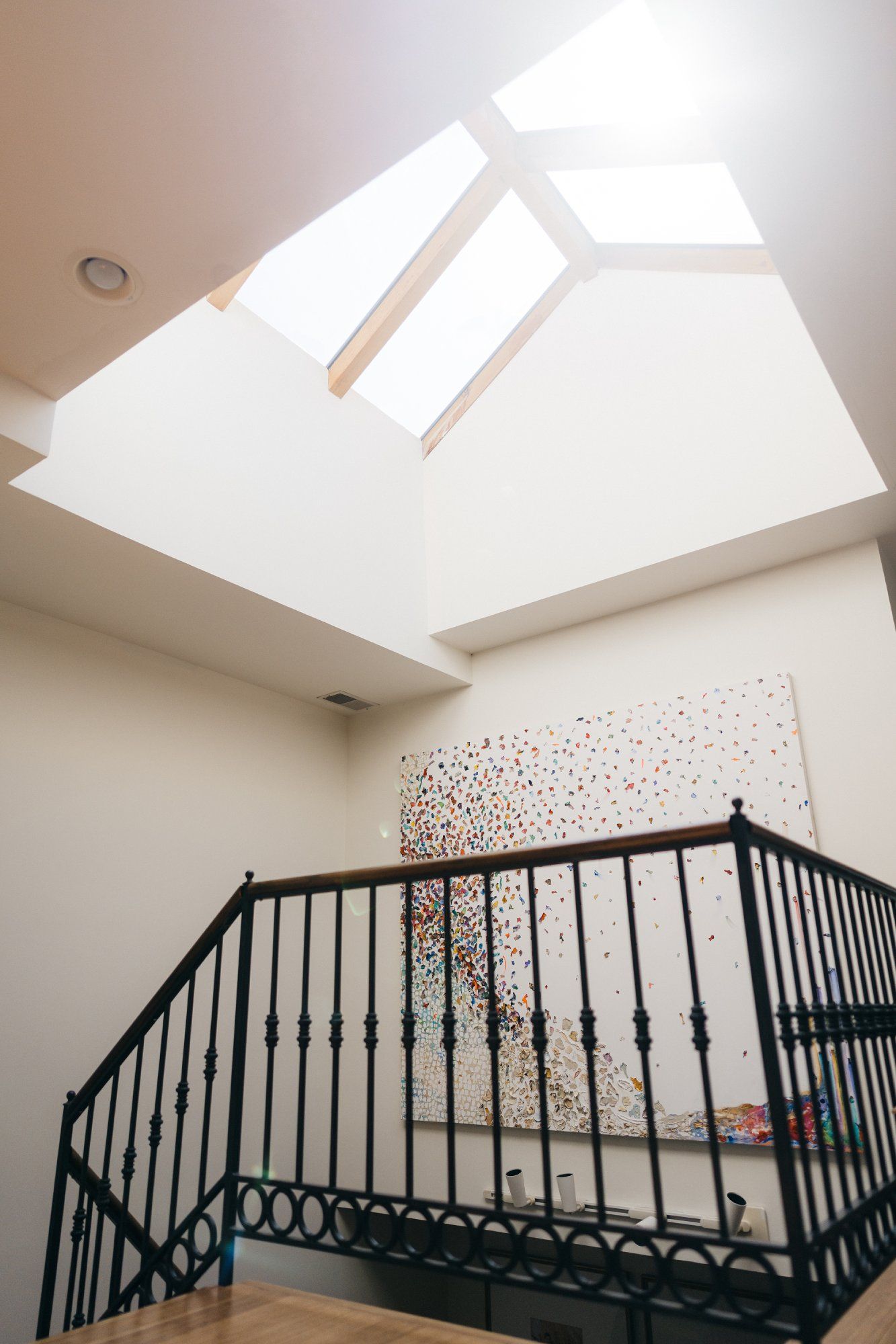 A modern interior with bright skylight, white walls, and colourful abstract wall art, complemented by a decorative black wrought iron railing