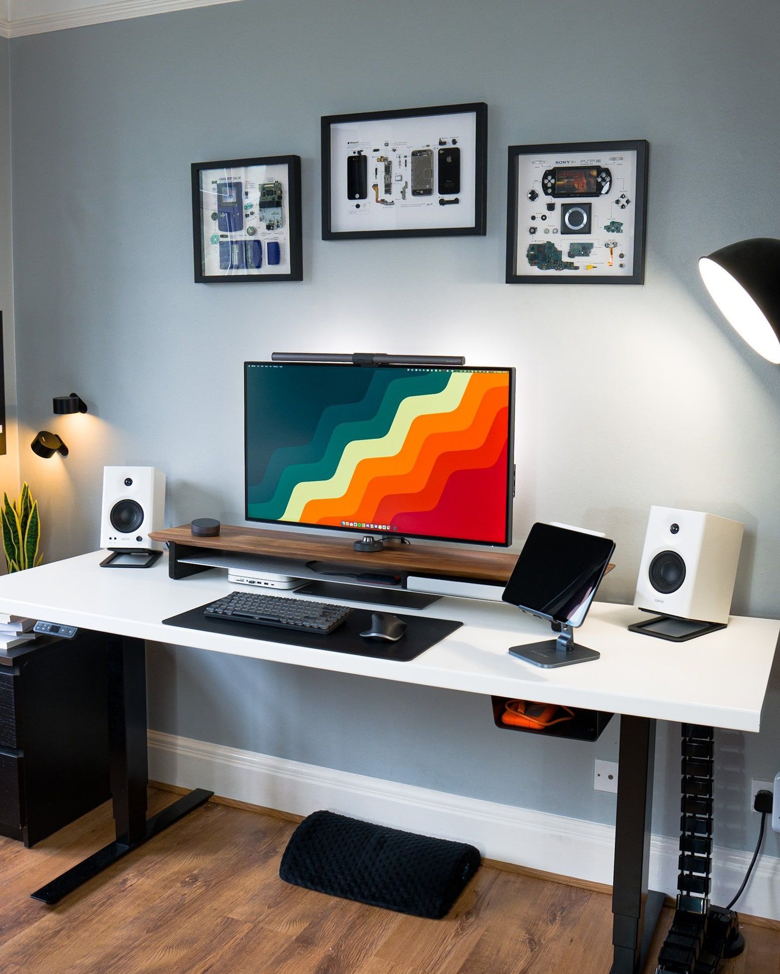 A home office desk setup with a white desk, colourful widescreen monitor, high-fidelity speakers, and a tablet stand, against a backdrop of grey walls with framed technical art