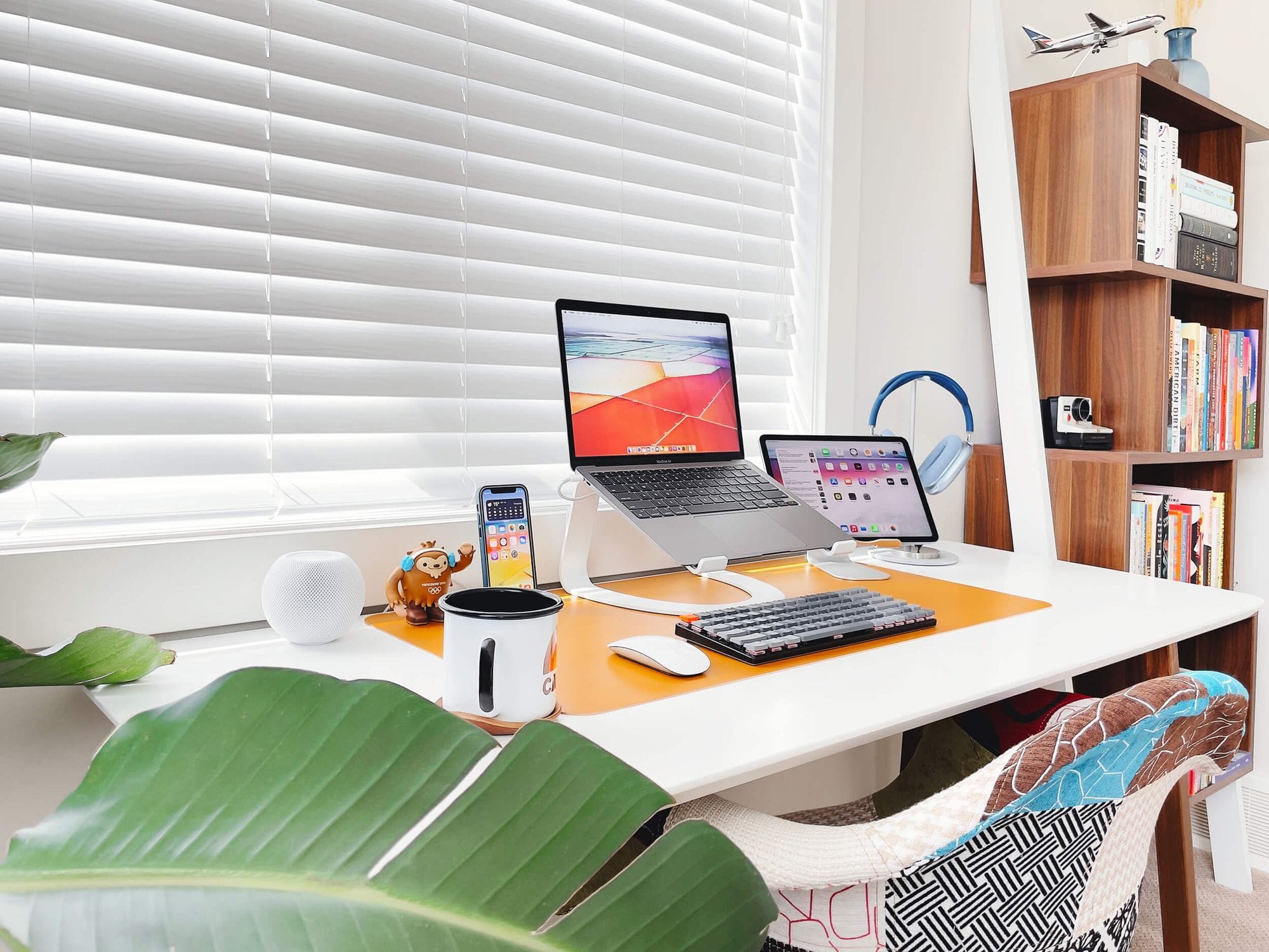 A cheerful home office space with a MacBook, iPad, iPhone on a charging dock, and a mechanical keyboard on a white desk mat, complete with a coffee mug, decorative figurine, HomePod, and cosy quilted chair, against a backdrop of white blinds and a bookshelf