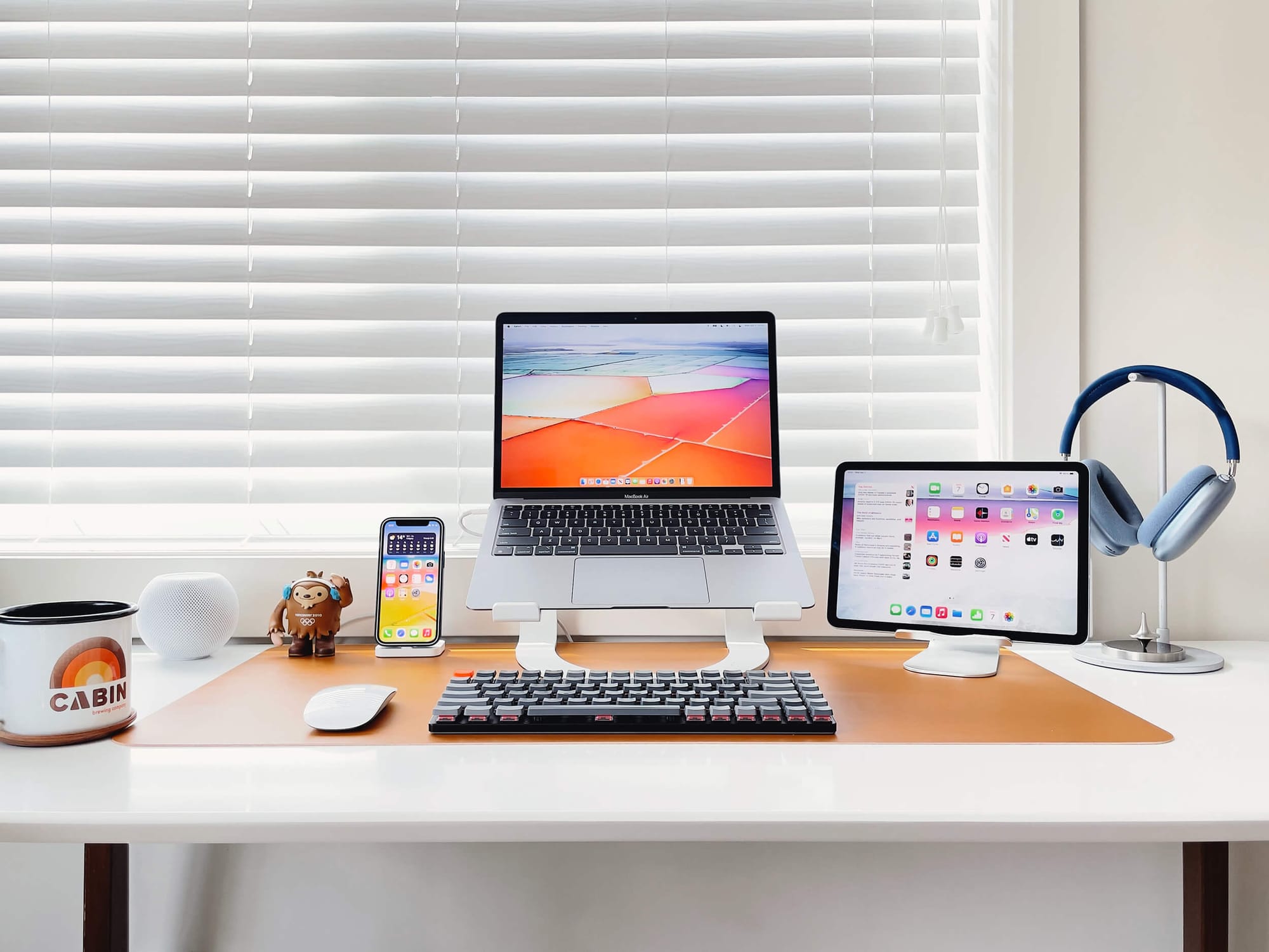 A bright and organised desk setup featuring an Apple MacBook on a stand, iPad with keyboard, iPhone, HomePod, mechanical keyboard, and headphones, all neatly arranged on a white desk against a window with white blinds