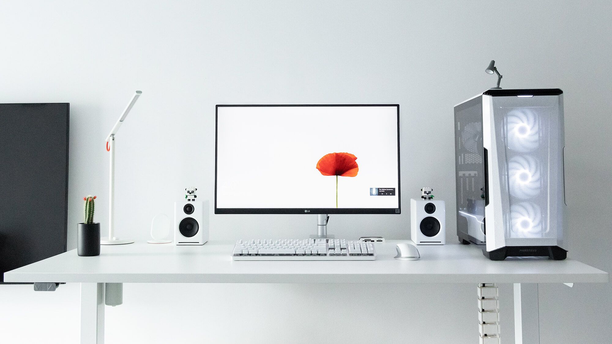 A minimalist cable-free desk setup with a white theme, featuring a sleek monitor with a poppy flower on the screen, a mechanical keyboard, a modern PC case with internal LED fans, stylish desktop speakers, a potted cactus, and a chic desk lamp, all against a clean grey backdrop