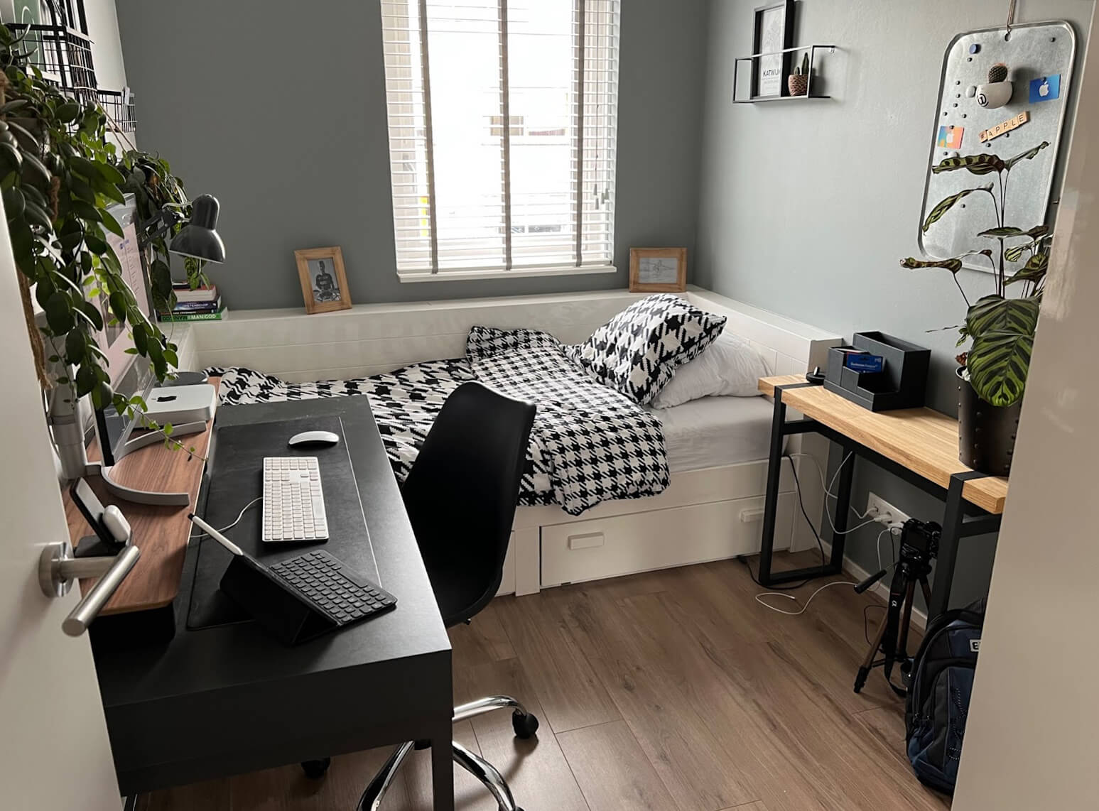 A neat, multifunctional room with a single bed covered in a black and white patterned comforter next to a window with blinds. In front of the bed is a black desk with a white keyboard, tablet, and lamp, complemented by a black office chair