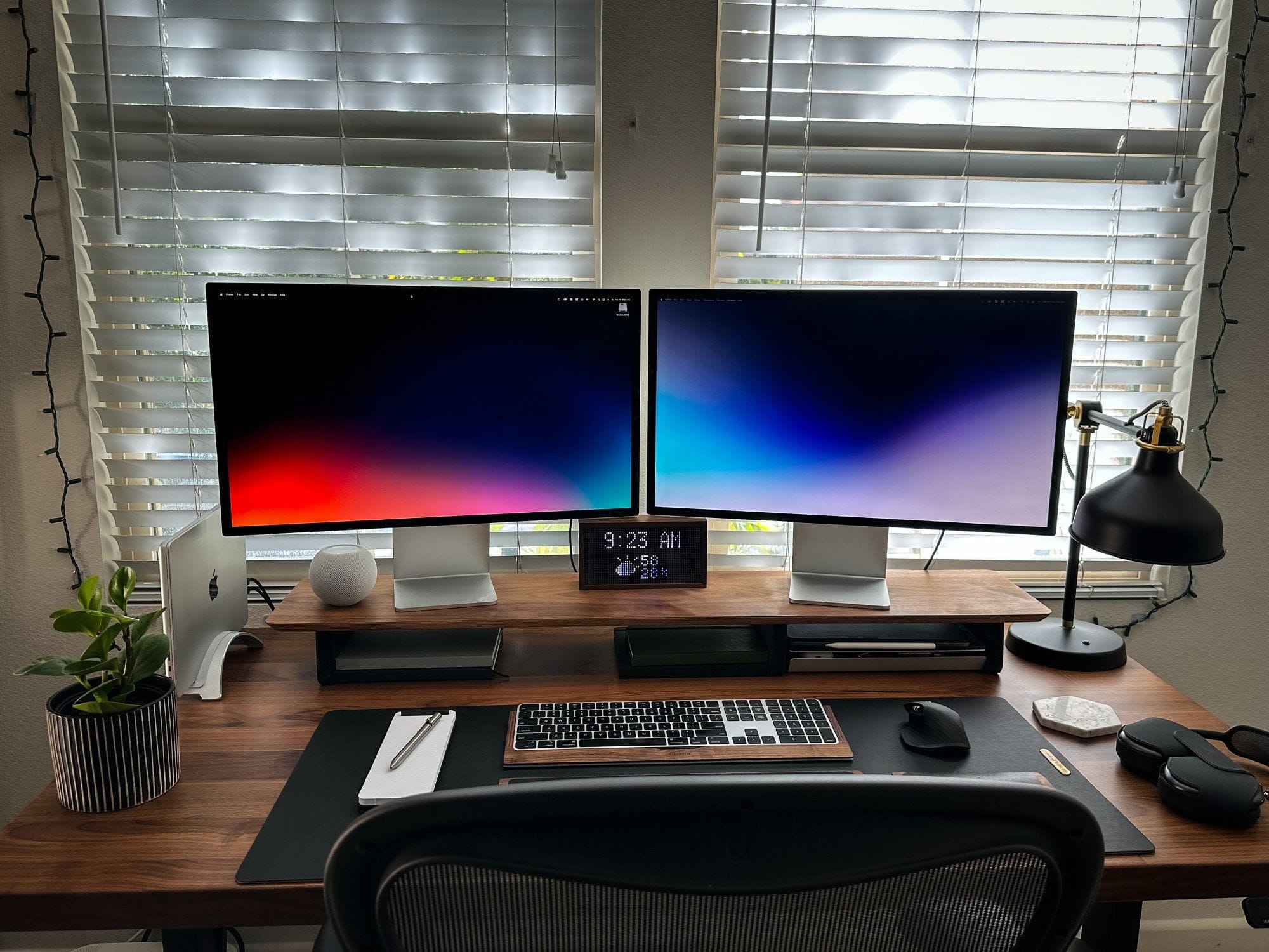 A well-organised dual monitor Apple desk setup with ambient lighting, featuring an iMac, MacBook Pro on a stand, a HomePod, desk plant, desk lamp, and various accessories, positioned in front of a window with Venetian blinds